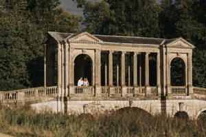 Elegant neoclassical stone colonnade with white pillars and archways in a park setting during golden hour.