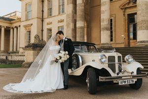 Elegant wedding couple shares romantic moment beside vintage cream Beauford car at grand historic building entrance.