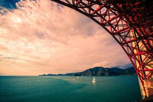 Sailboats float beneath red Golden Gate Bridge arch with dramatic sunset sky and turquoise San Francisco Bay waters.