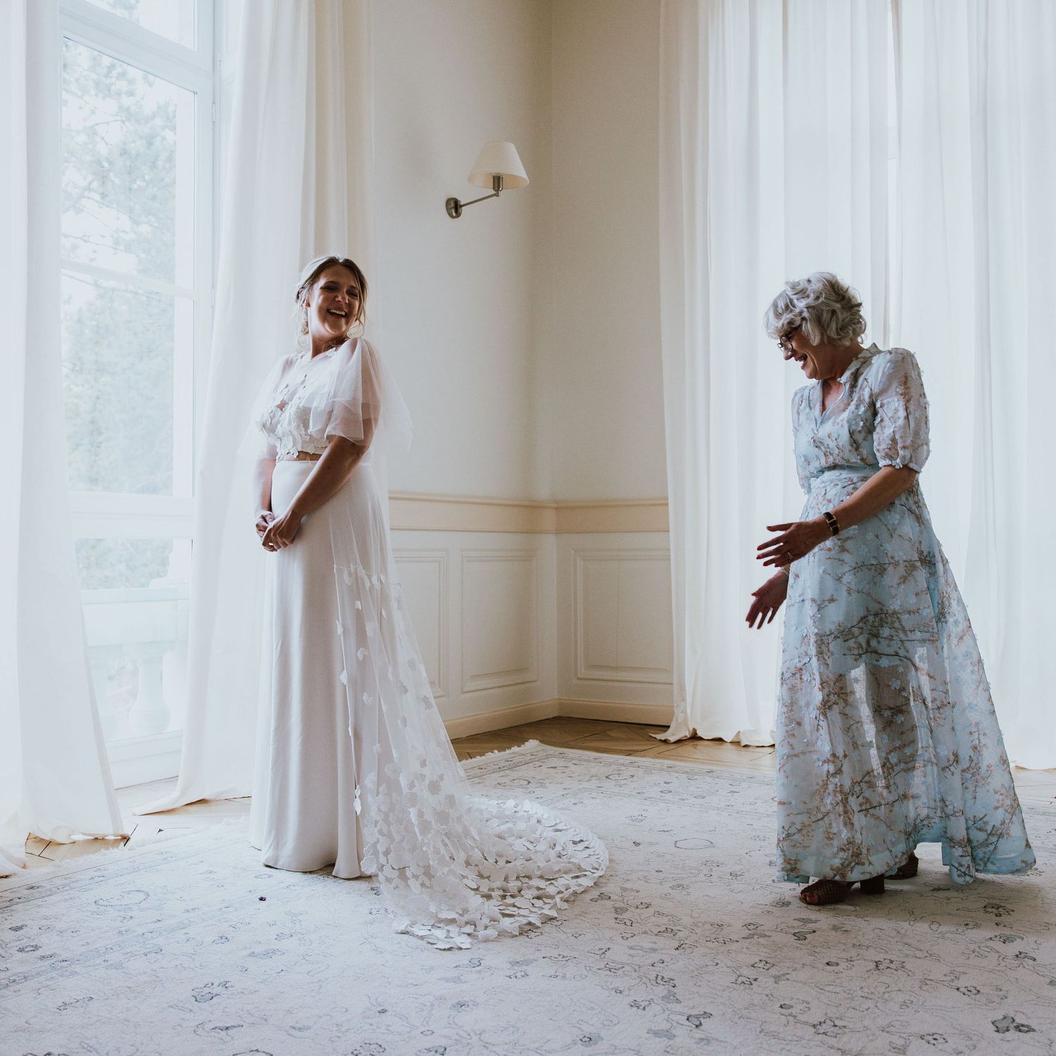 Two people stand in an elegant white room wearing long lace dresses during a special moment.