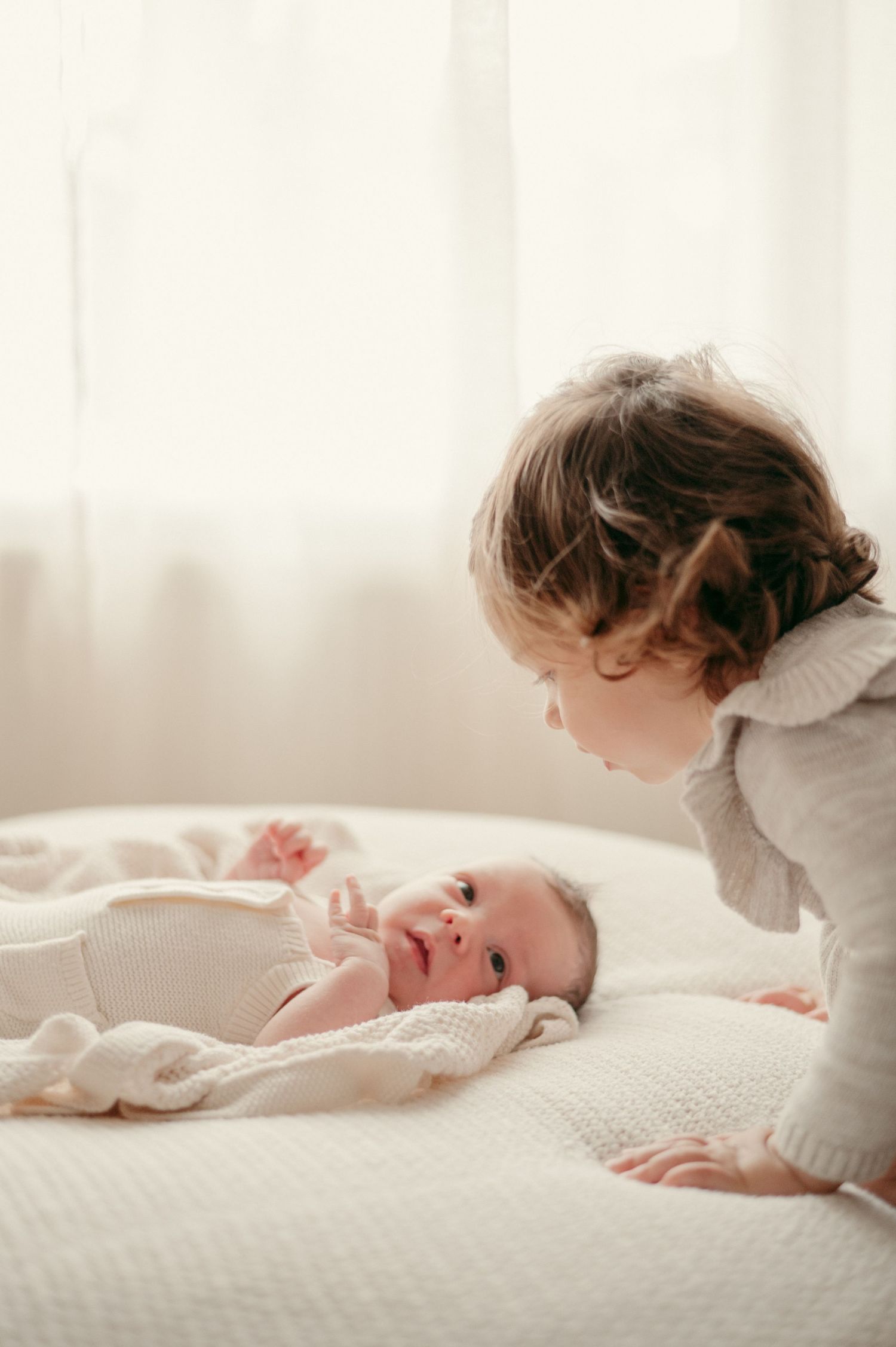 Touching moment between siblings as an older child looks at a newborn baby lying on white bedding.