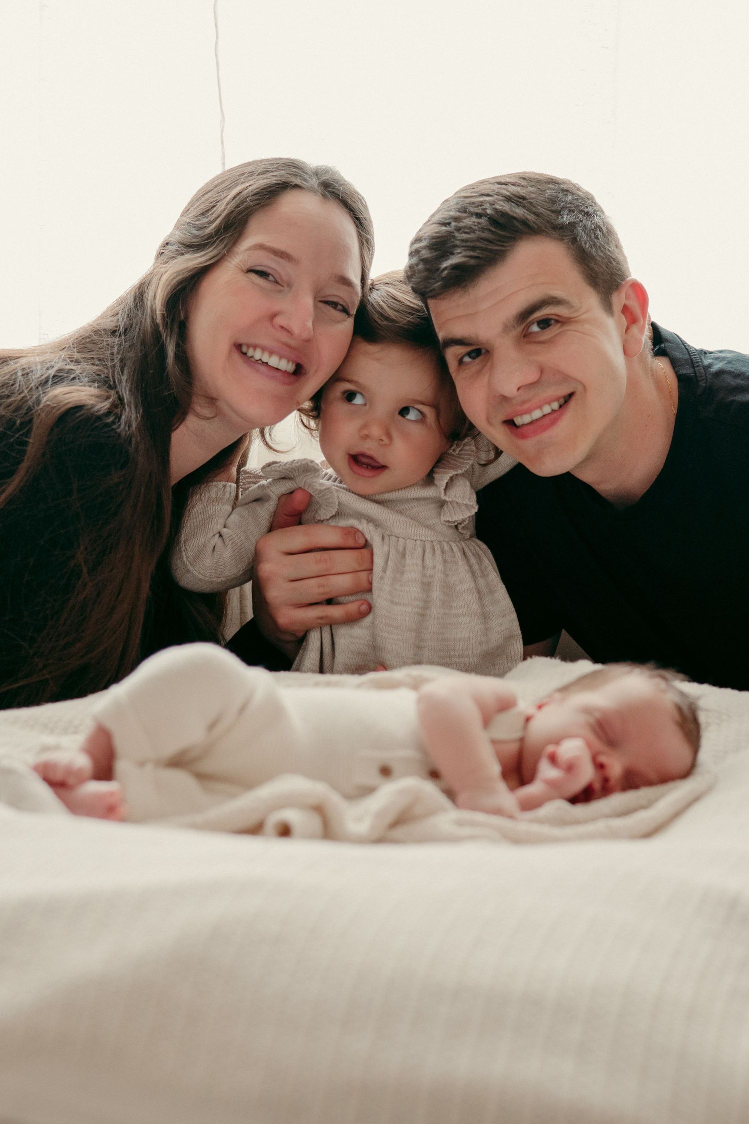 Family portrait with newborn baby sleeping peacefully on white bedding.