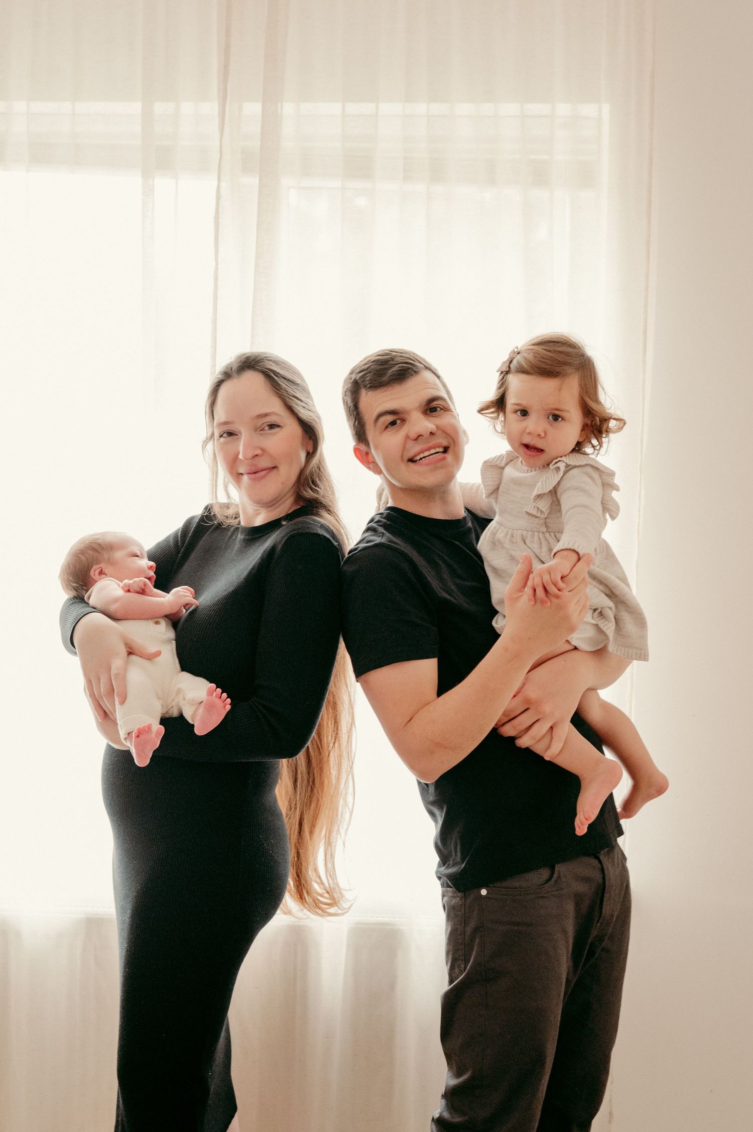 Indoor family portrait with parents holding their children against a bright window background.