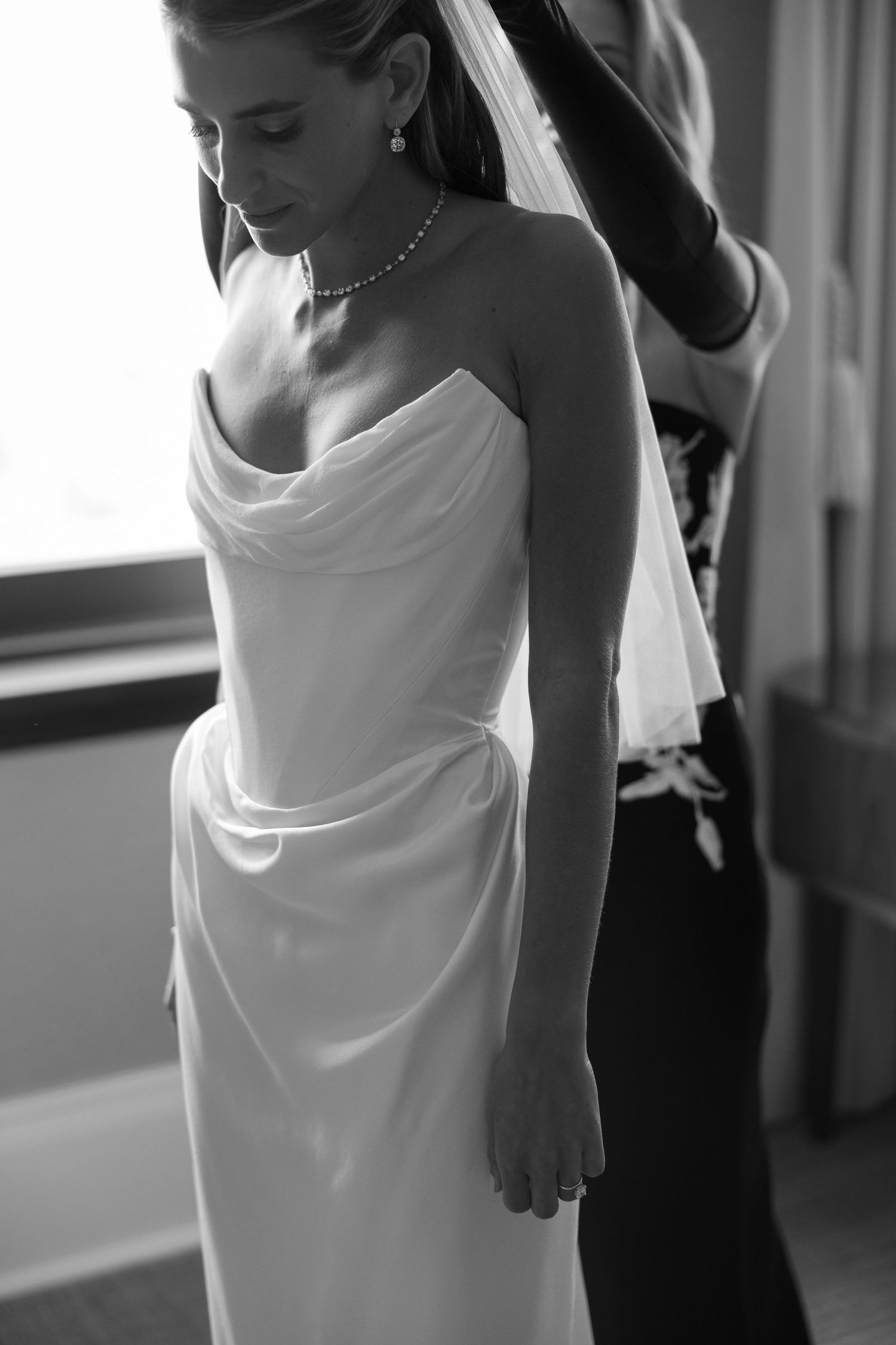 Black and white photo of a bride in an elegant strapless draped wedding gown with flowing veil and delicate necklace.