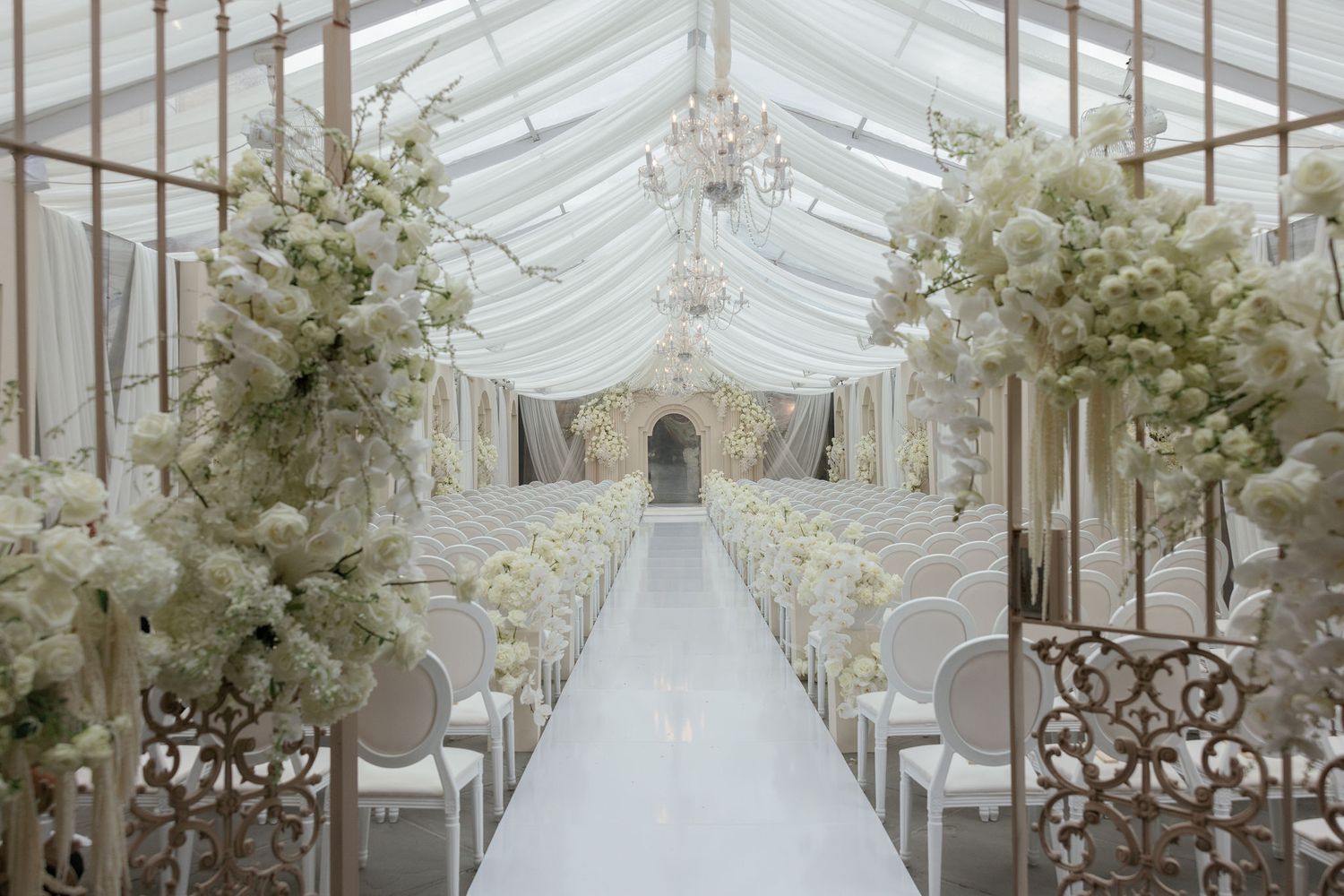 Elegant glass-covered wedding venue with white floral arrangements and crystal chandeliers along a pristine white aisle.
