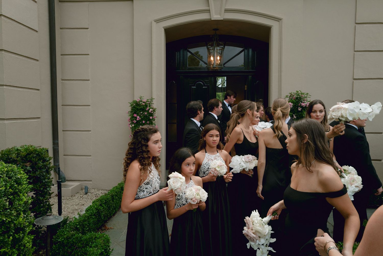 Wedding party gathers on church steps as bridesmaids in black dresses hold white bouquets during outdoor group photo.