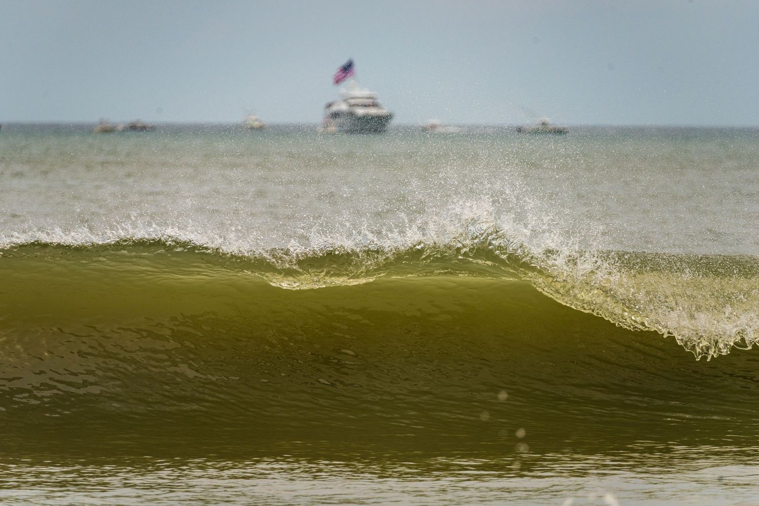 A green ocean wave crests with spray as a boat sits on the horizon against a blue grey sky.