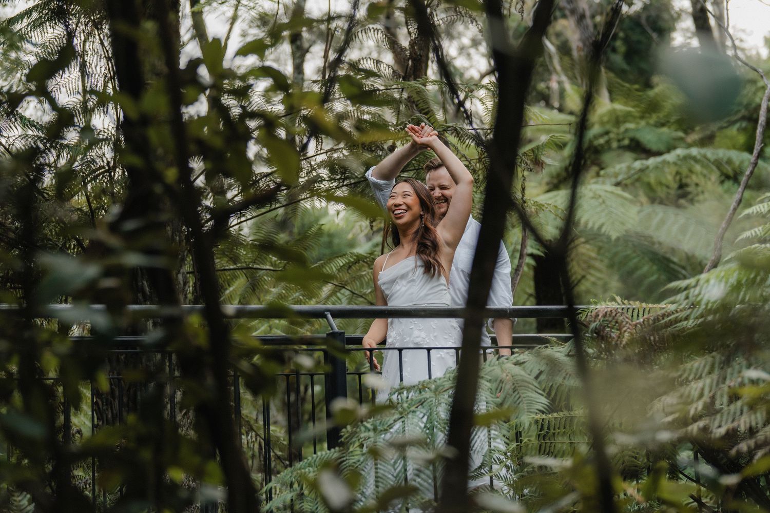 A couple embraces while taking a selfie on a rustic bridge surrounded by lush forest ferns and greenery.