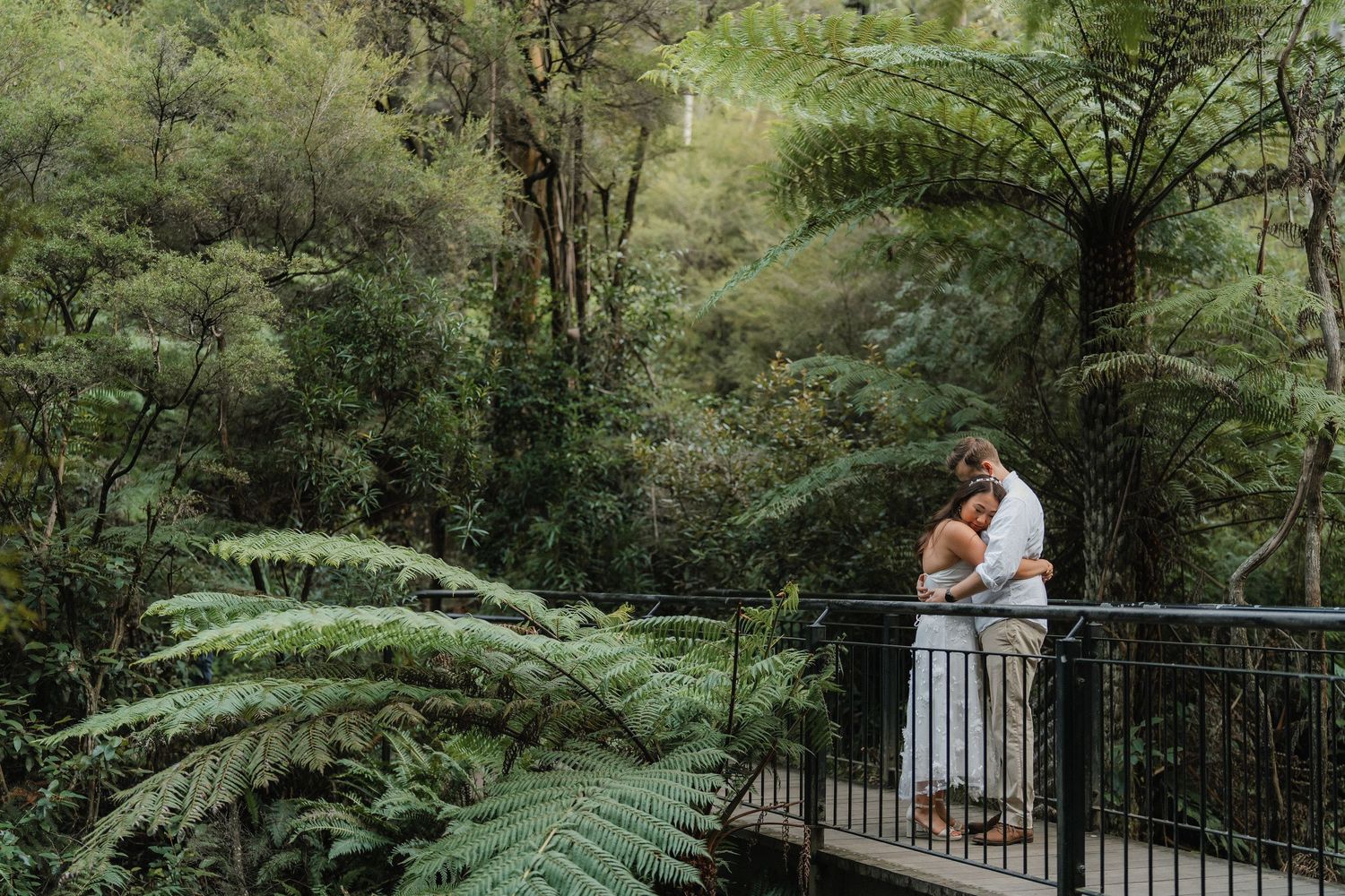 A romantic moment between two people standing on a bridge walkway amidst tropical forest and ferns.