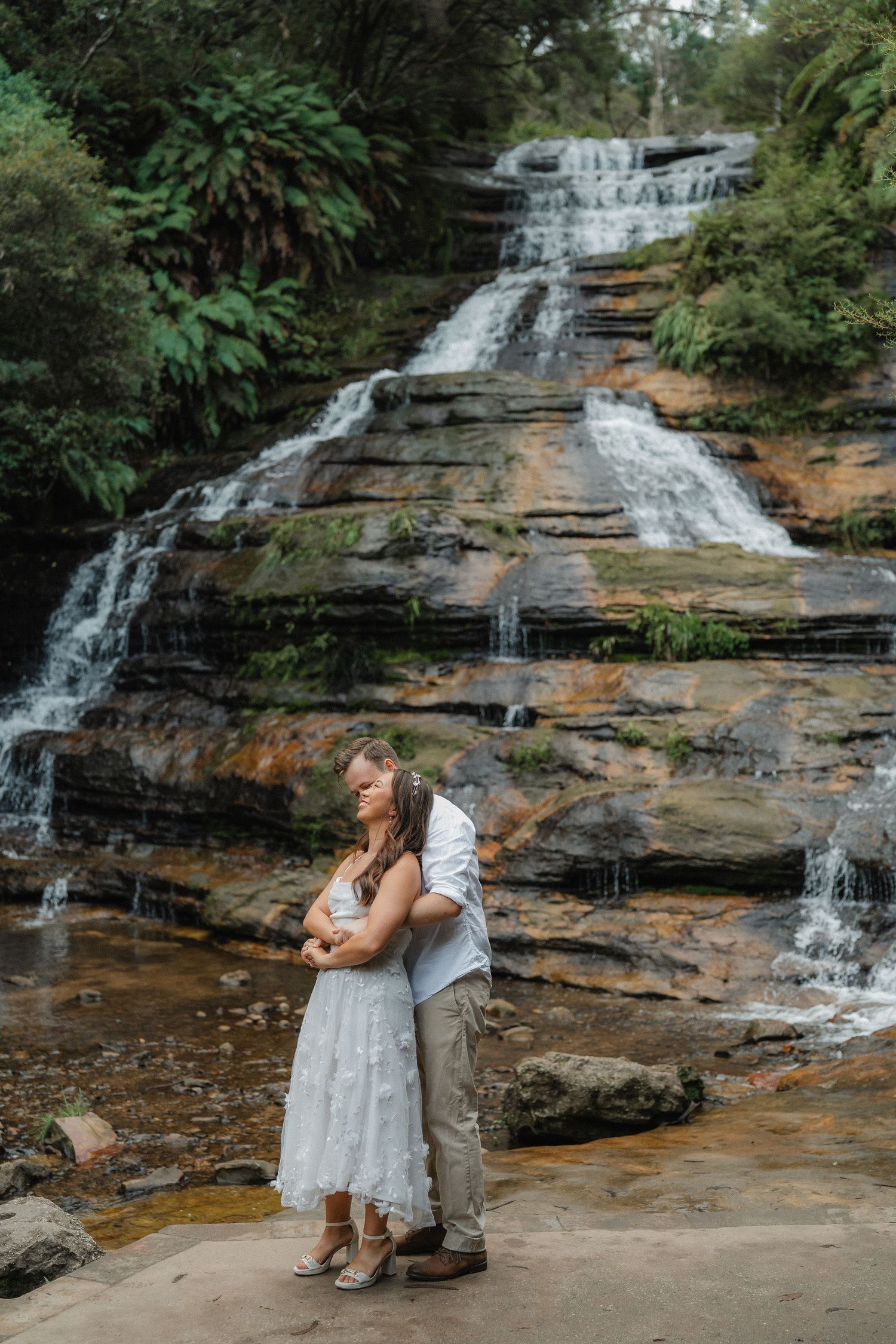 A couple embraces in front of a multi-tiered waterfall cascading down rocky cliffs.