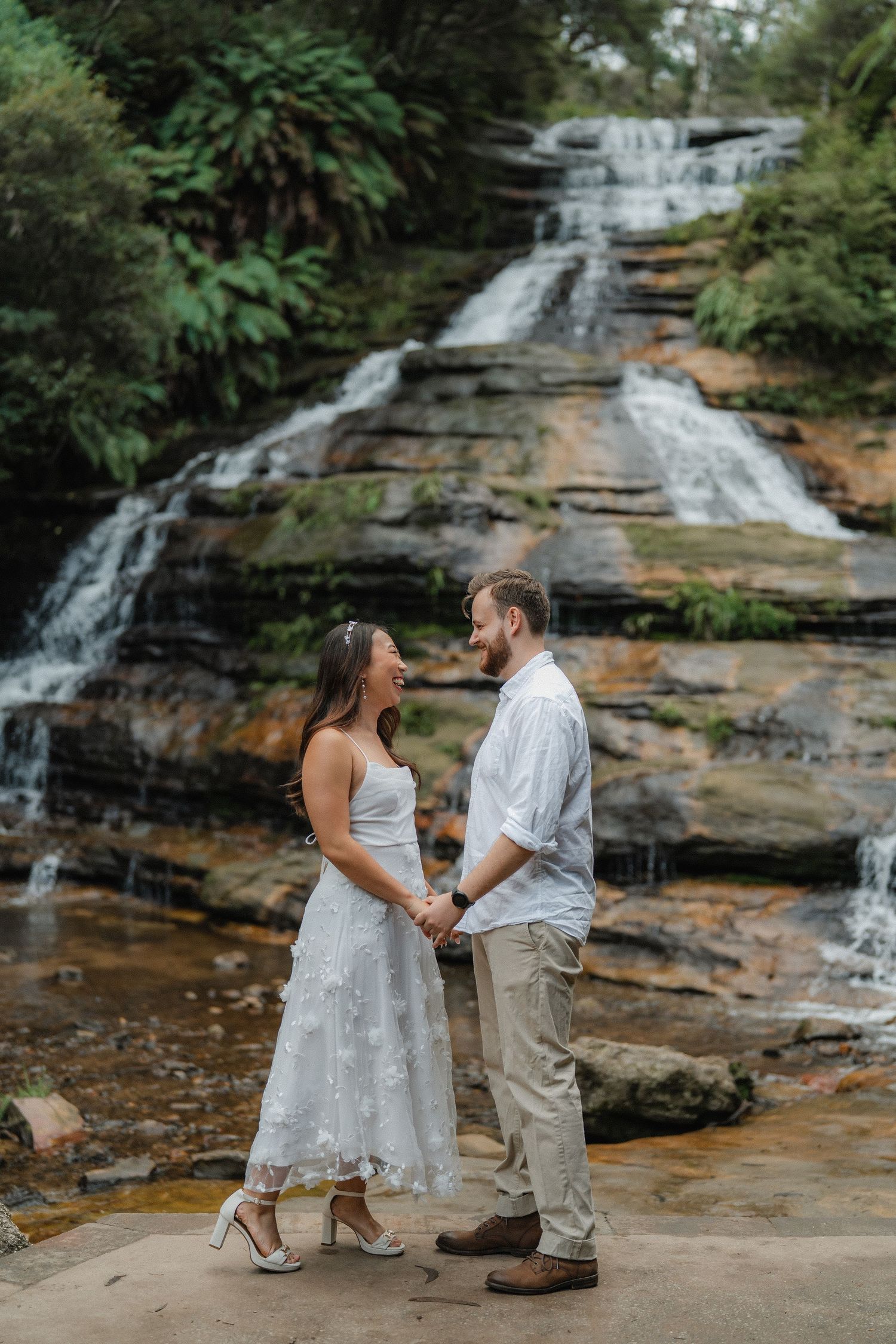 A couple stands together on a rock platform with a beautiful cascading waterfall behind them.