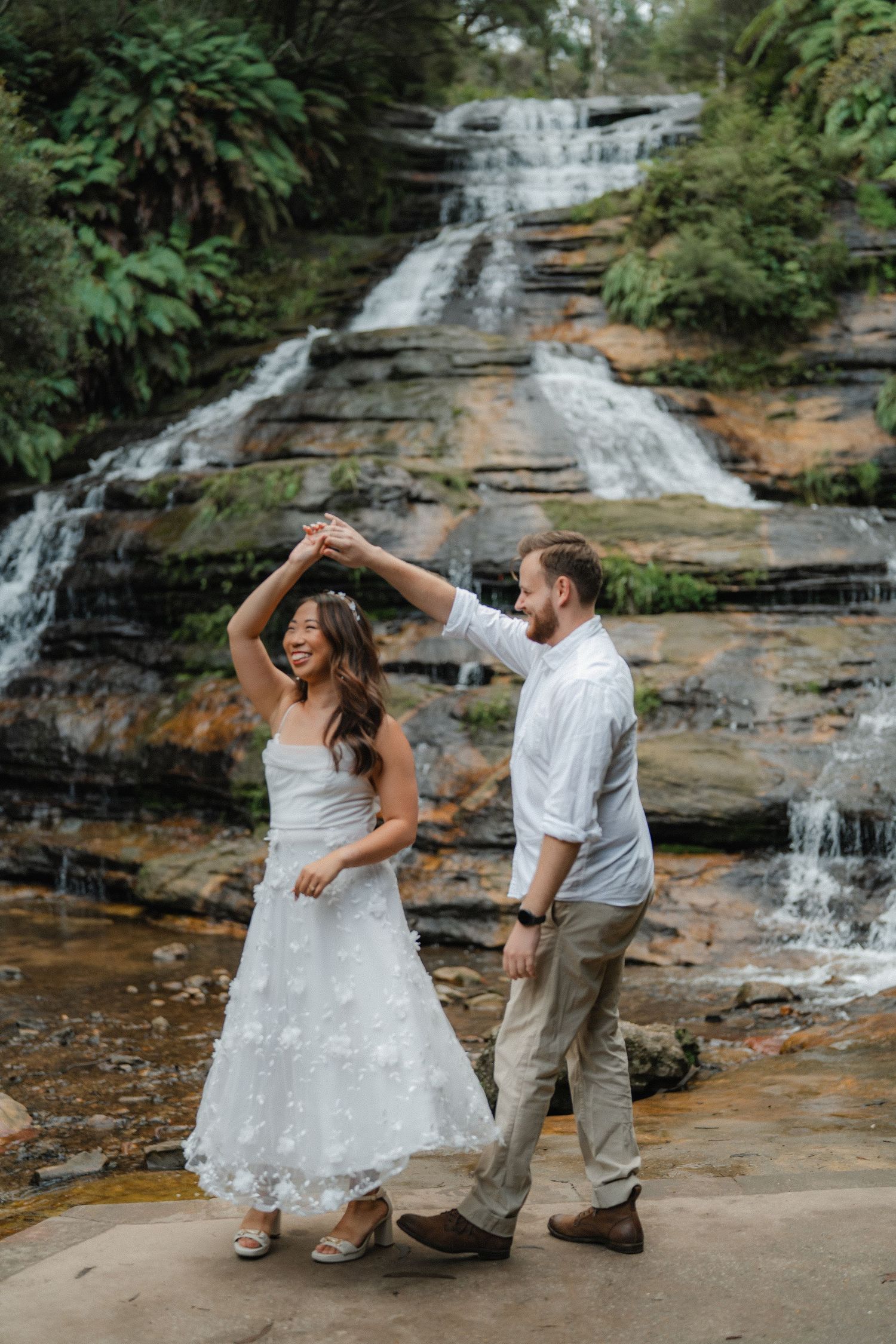 A couple dances together on a rocky platform with a cascading waterfall as their backdrop.