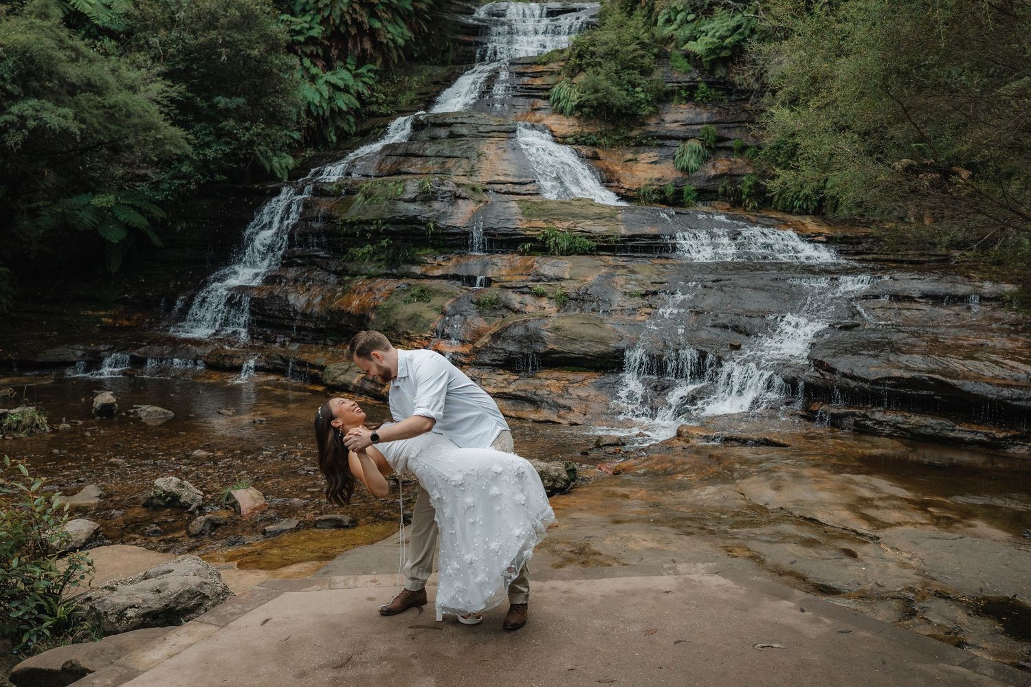 A couple shares a romantic moment on rocks in front of a scenic multi-tiered waterfall.