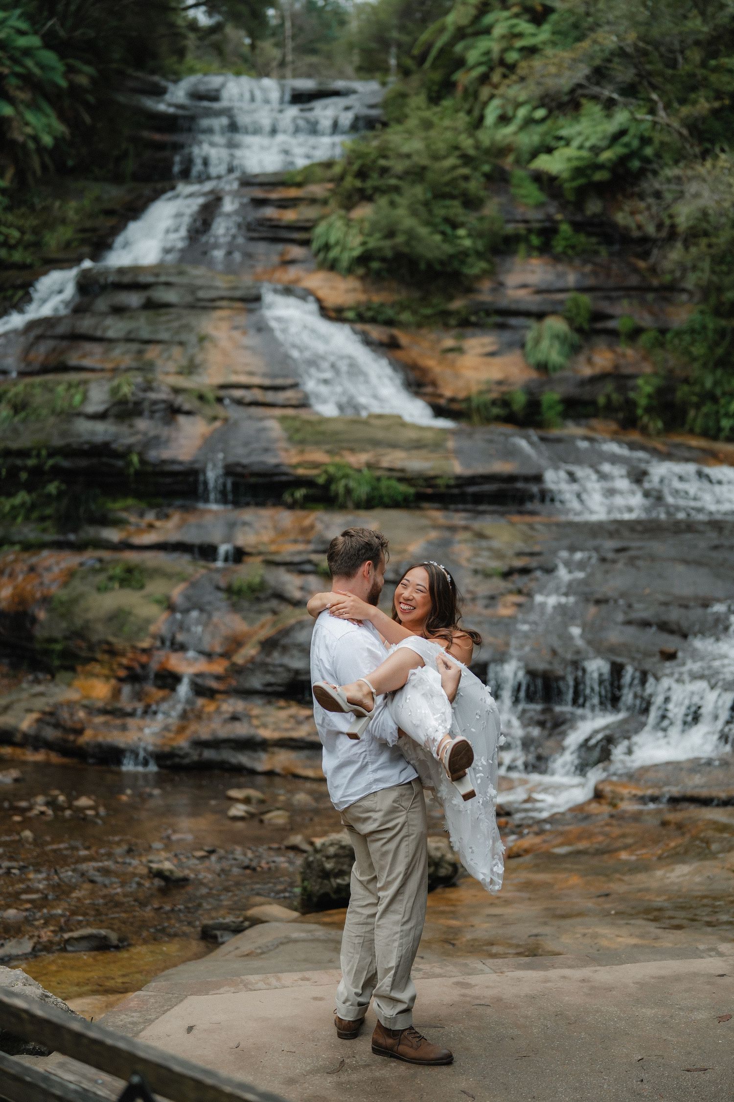 A playful moment captured as a couple spins in front of a scenic forest waterfall.