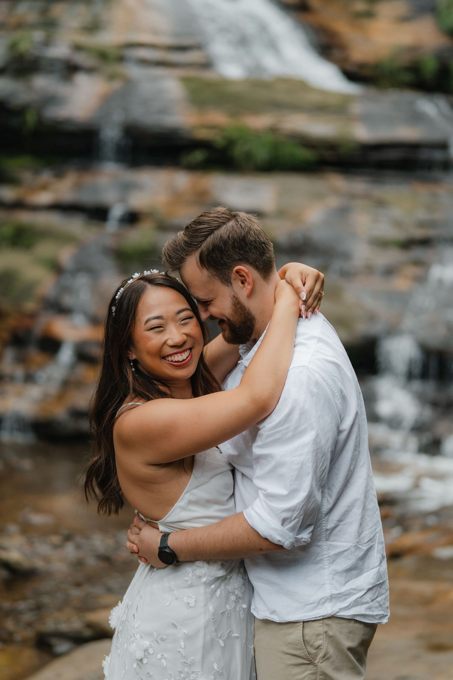 Two people share an intimate embrace in front of a scenic waterfall with rocky cliffs.
