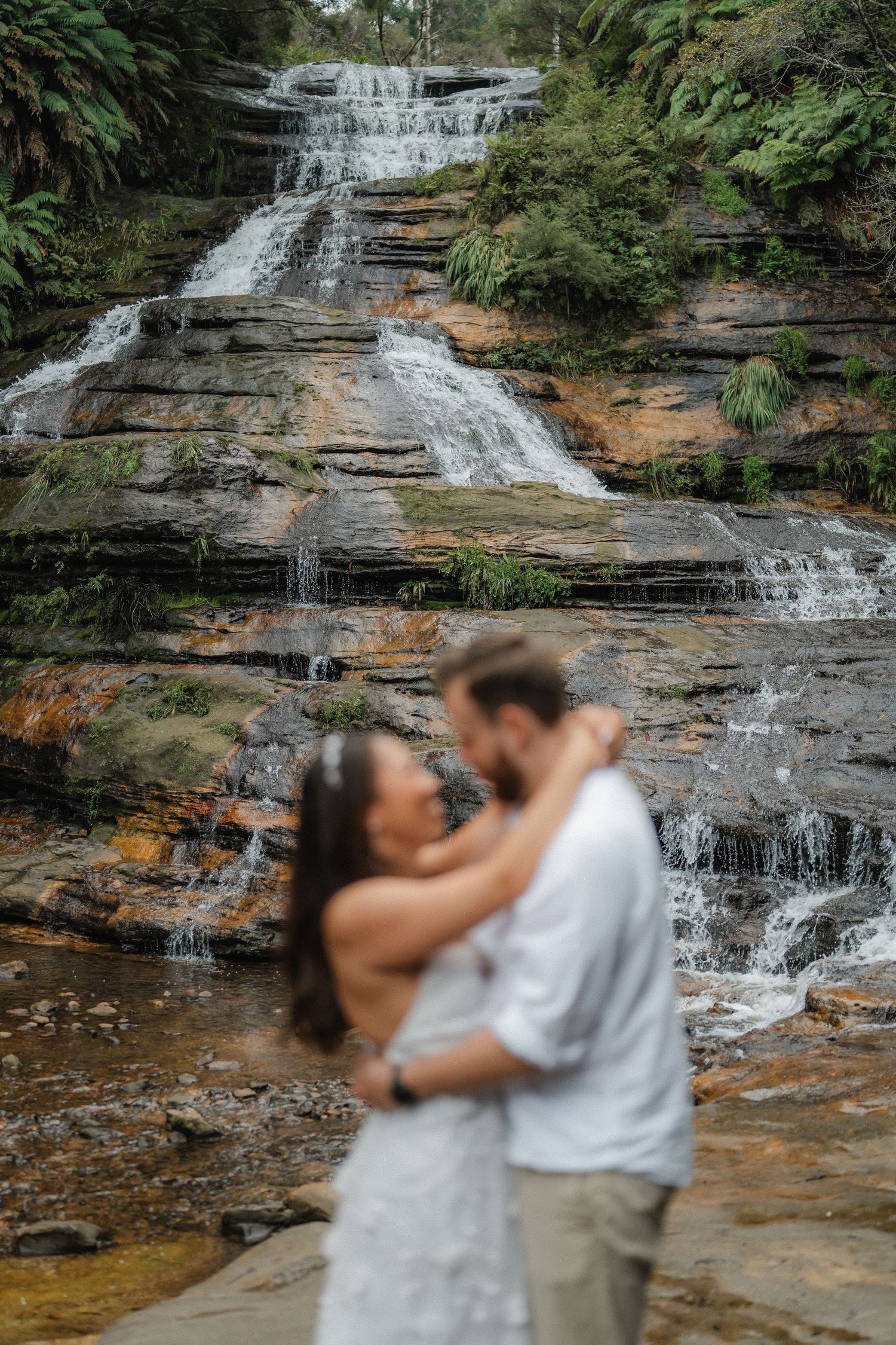 Couple embraces in front of cascading waterfall with rustic rock formations and lush greenery.