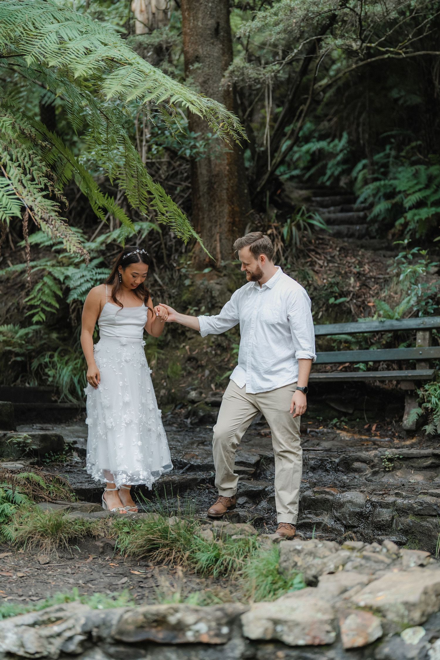 A couple shares a tender moment on stone steps surrounded by ferns and natural scenery.
