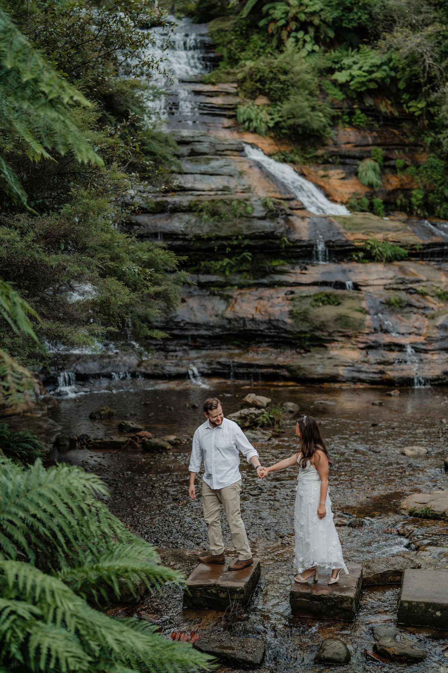 Couple shares tender moment by scenic waterfall with ferns and rocky terrain.
