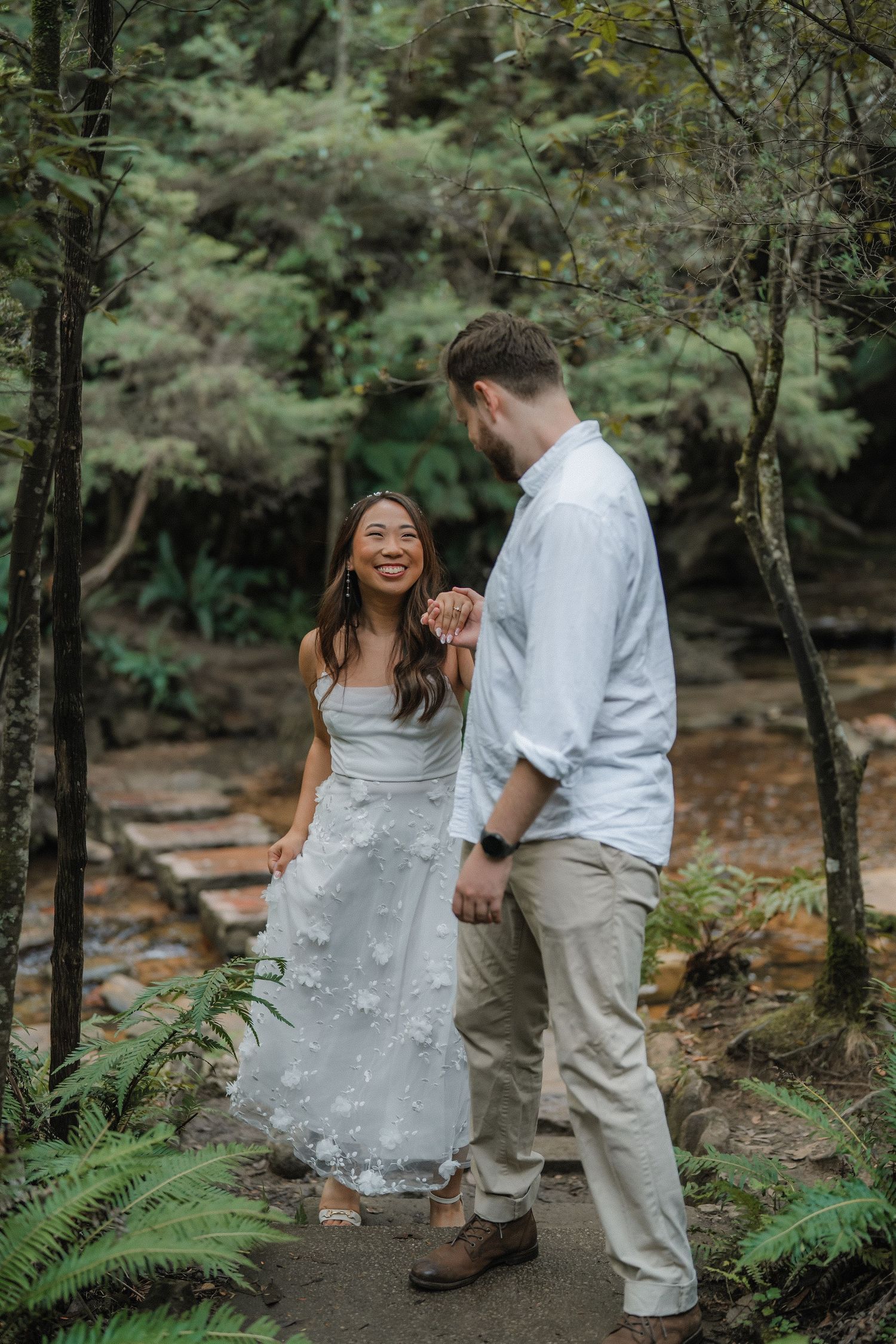 A couple shares a joyful moment together on a forest trail surrounded by ferns and trees.
