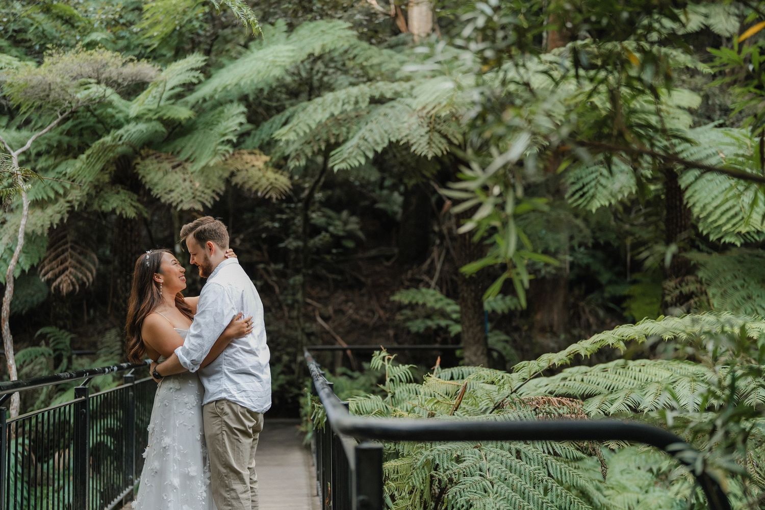A couple shares a tender moment on a forest walkway surrounded by lush ferns and greenery.