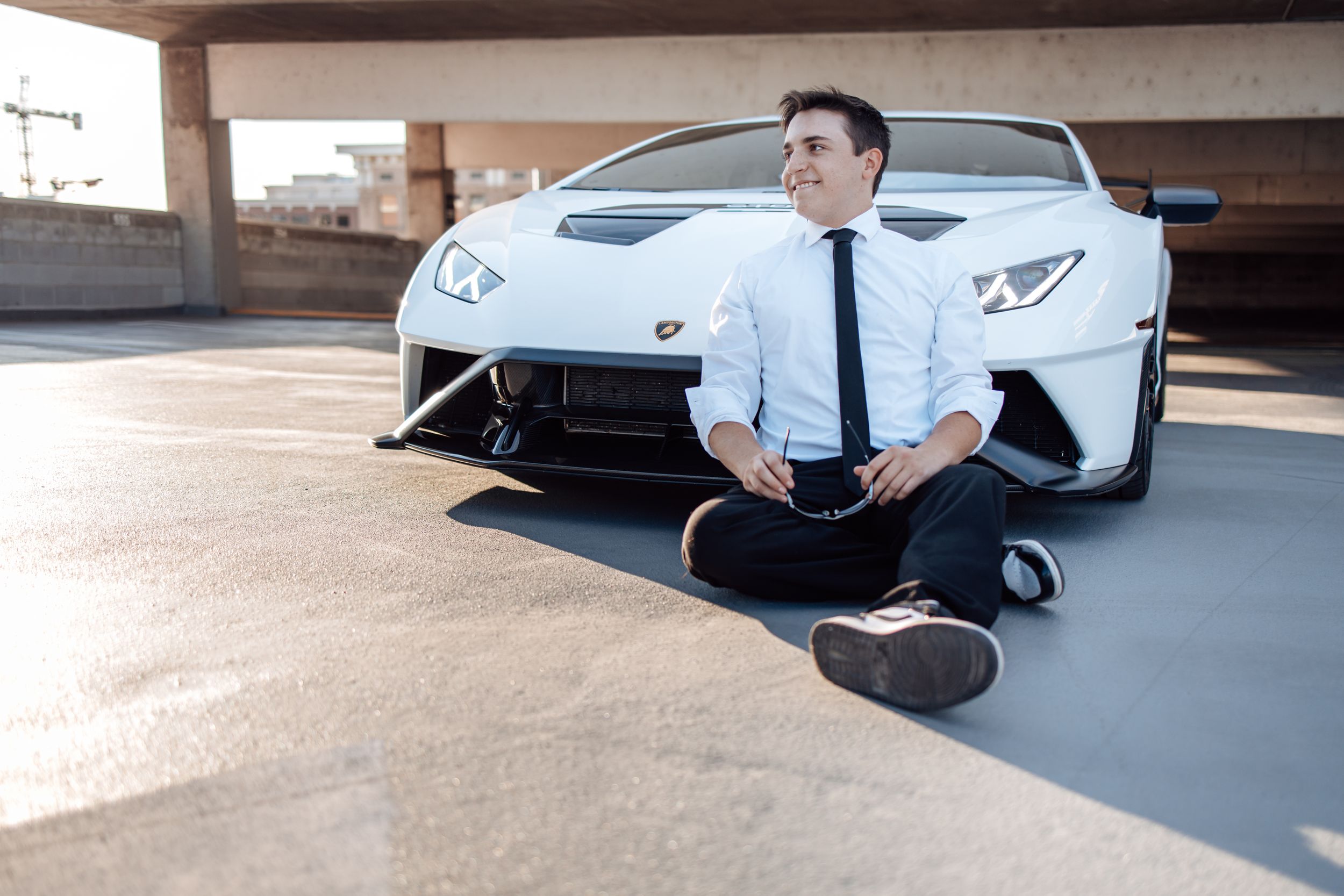 A white Lamborghini sports car parked in a concrete garage with someone in business attire sitting cross-legged in front.