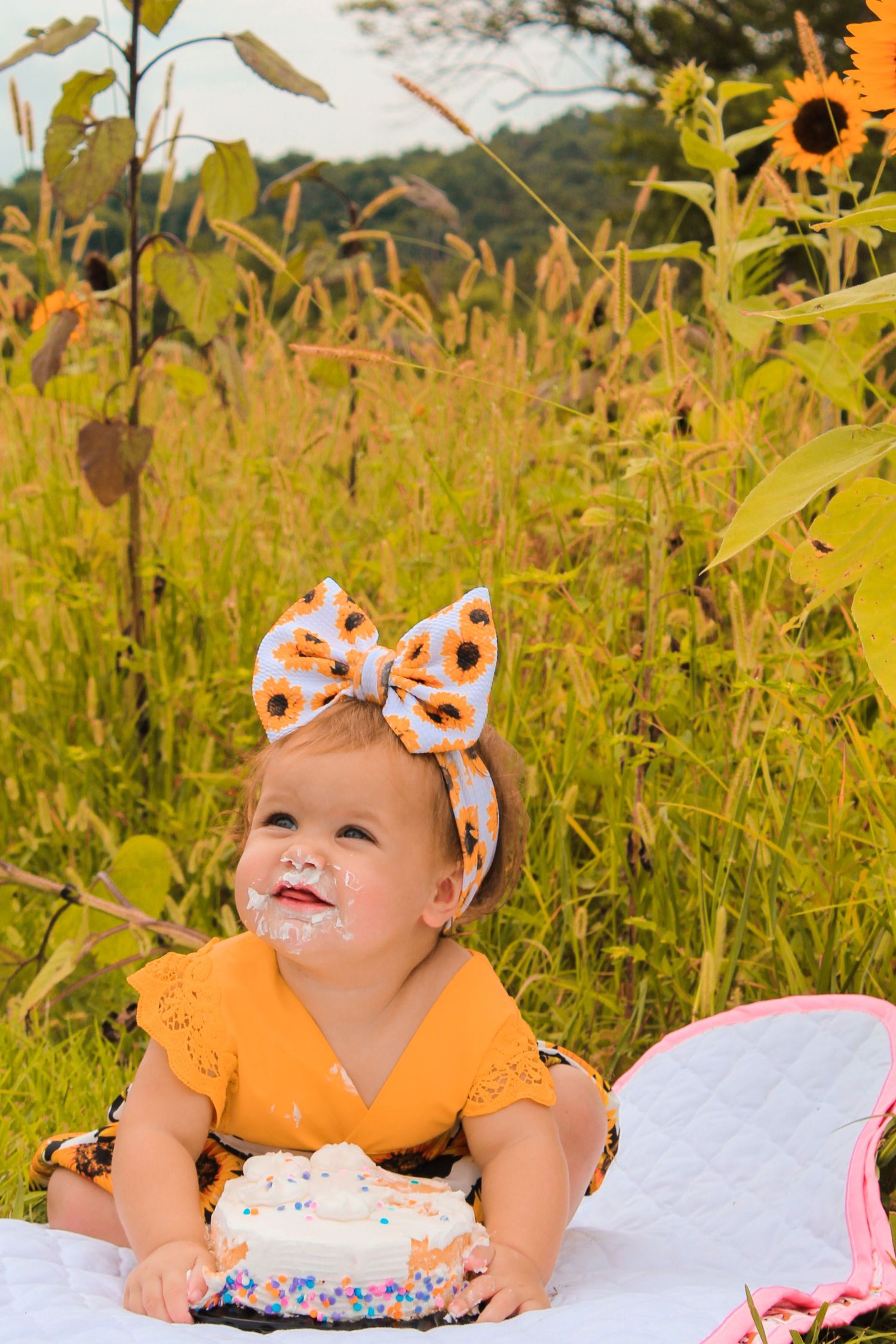 A small child wearing an orange top and patterned bow sits on a blanket with a cake against a backdrop of tall grass and sunflowers.