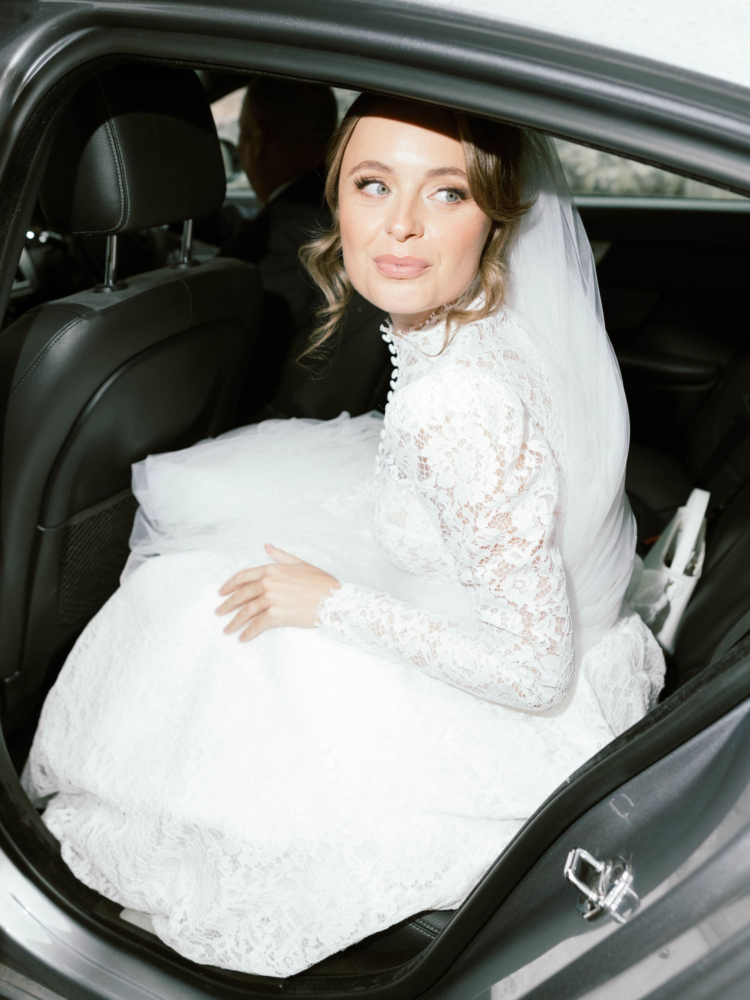 Elegant bride in a long-sleeved lace wedding dress sits gracefully in the backseat of a luxury car.