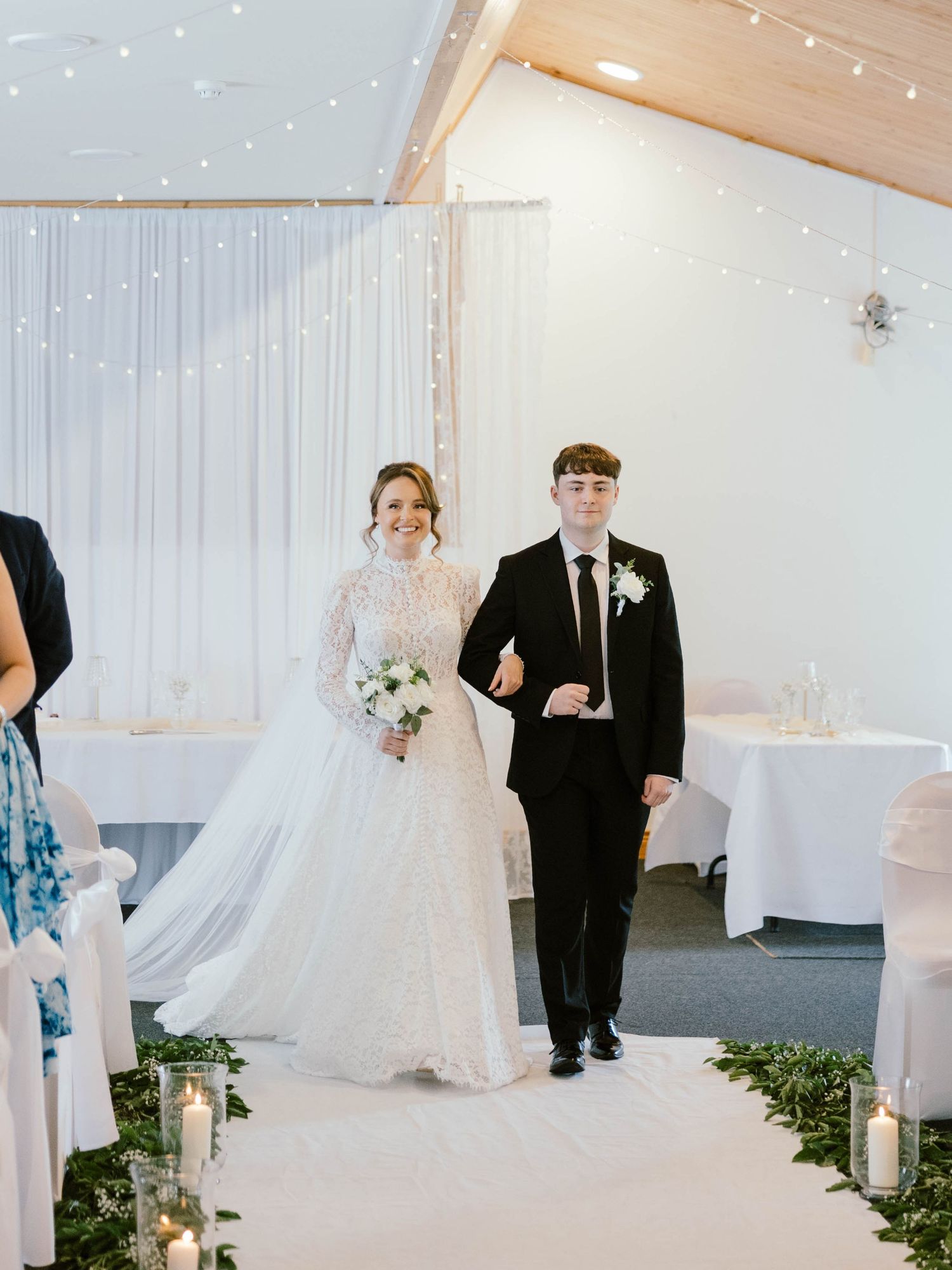 A couple walks down the aisle at their winter wedding ceremony with twinkle lights and white drapery decorating the venue.