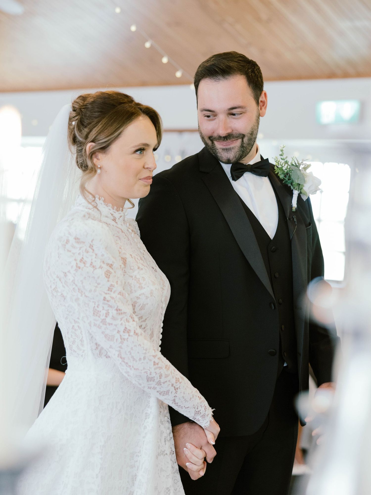 A bride in a white lace gown and groom in a black tuxedo share a tender moment during their wedding ceremony.