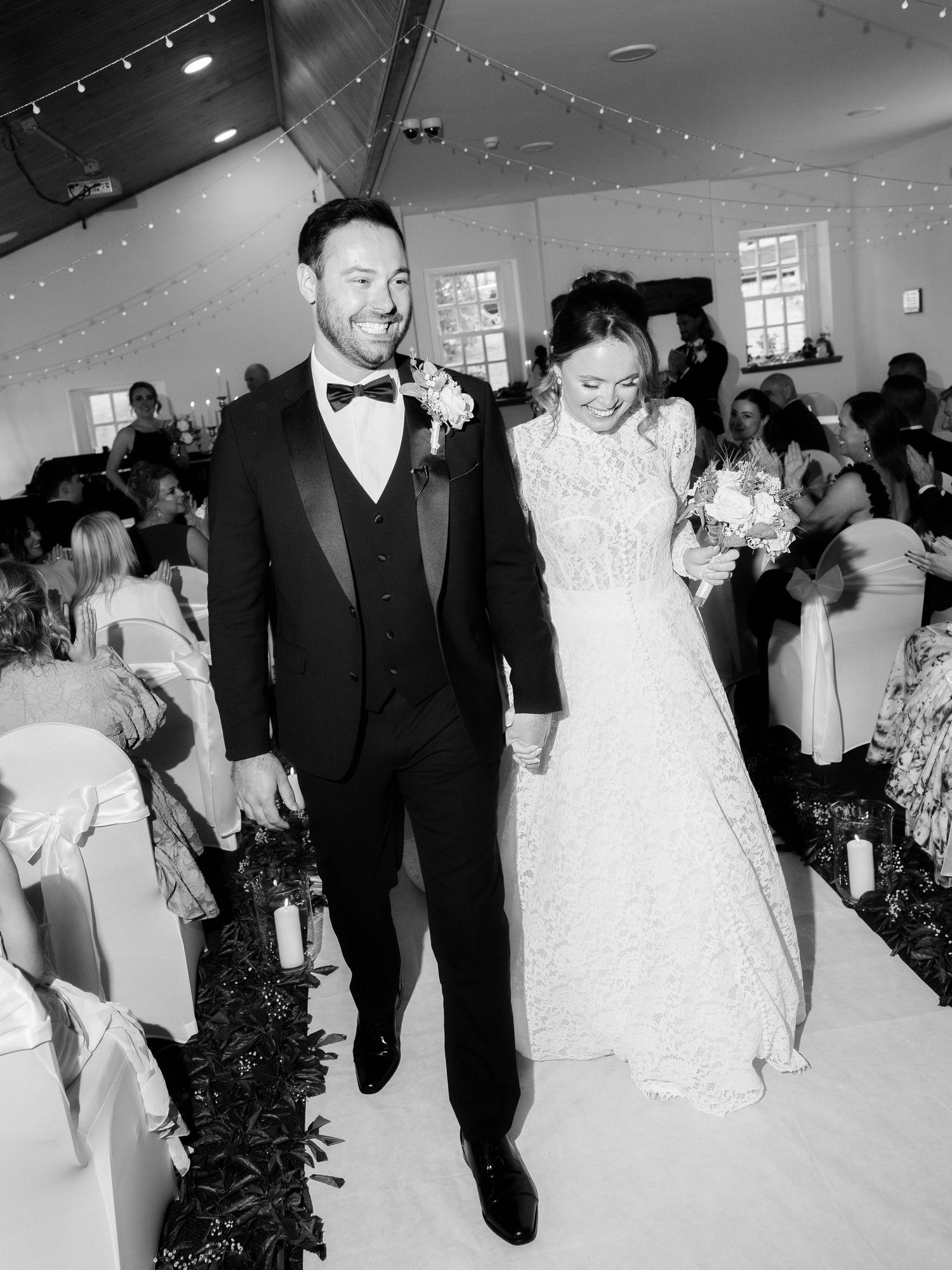 A newlywed couple walks down the aisle after their wedding ceremony in a church decorated with string lights.