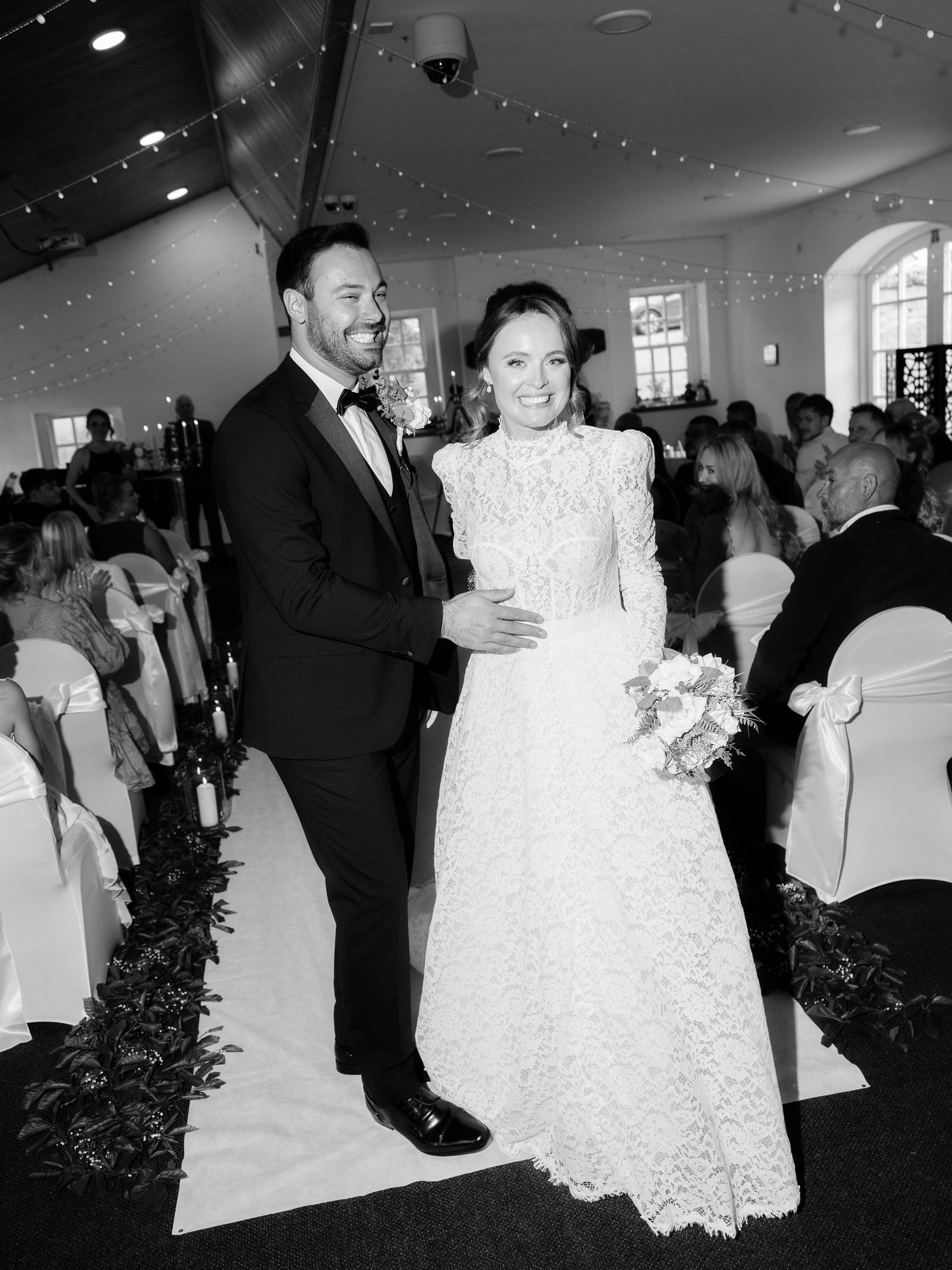 A wedding couple stands together on their special day in an elegant venue with twinkling lights and white decor.
