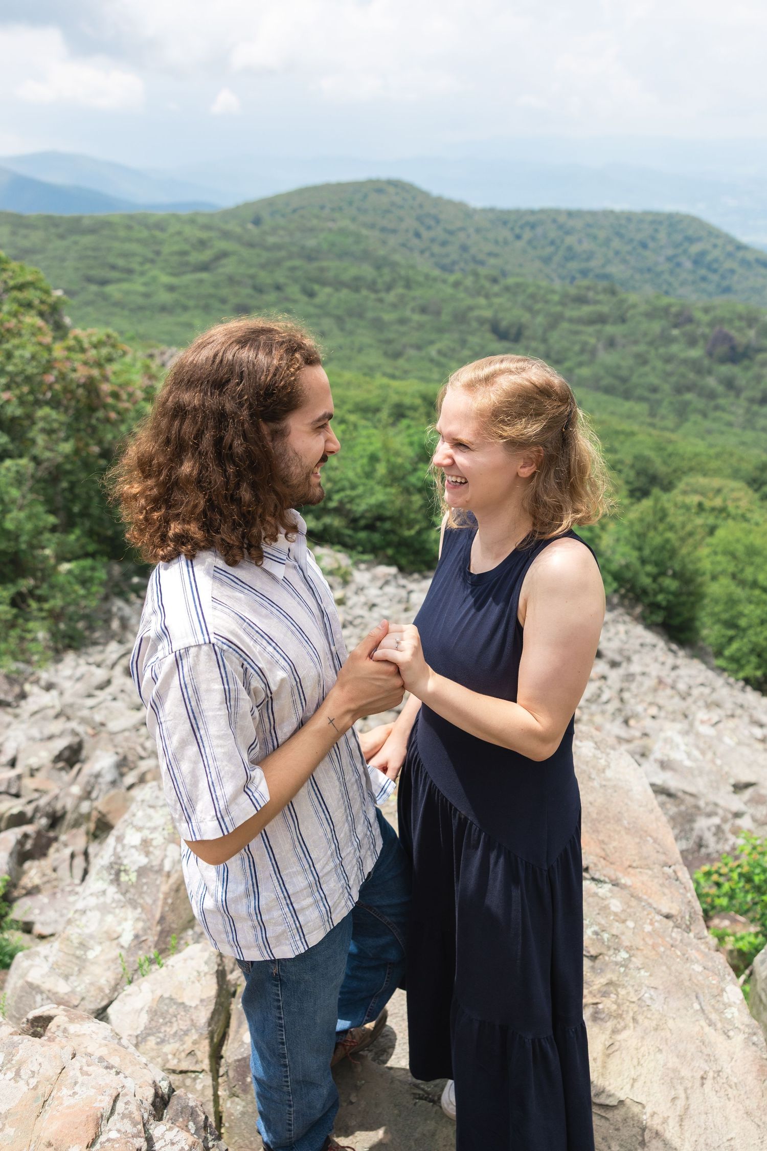 A couple shares a joyful moment while standing on rocky terrain with green mountains and forest in the background.