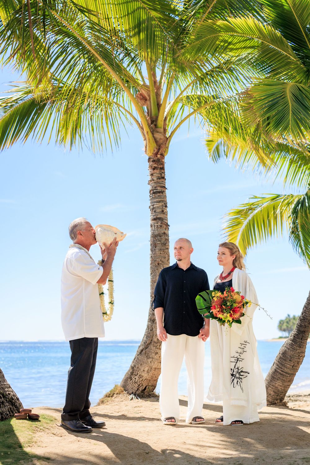 Couple exchanges vows in tropical beach wedding ceremony under palm trees with officiant blowing conch shell in Fiji.