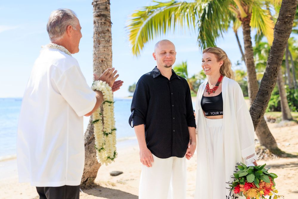 Couple exchanges vows on tropical beach wedding ceremony with officiant holding leis under palm trees.