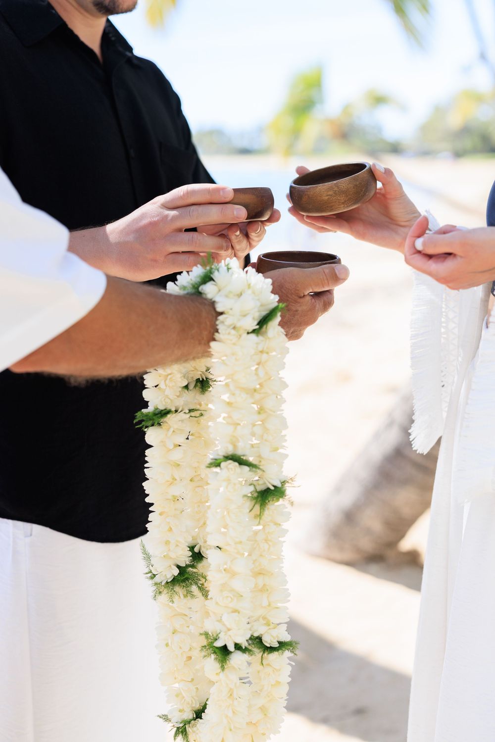 Couple exchanging floral leis and wooden bowls during a Hawaiian beach wedding ceremony.