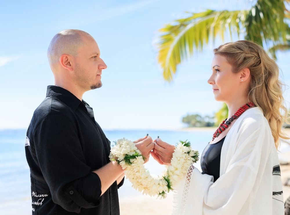 Couple exchanging flower leis during a beach wedding ceremony in Hawaii with palm trees and ocean in background.
