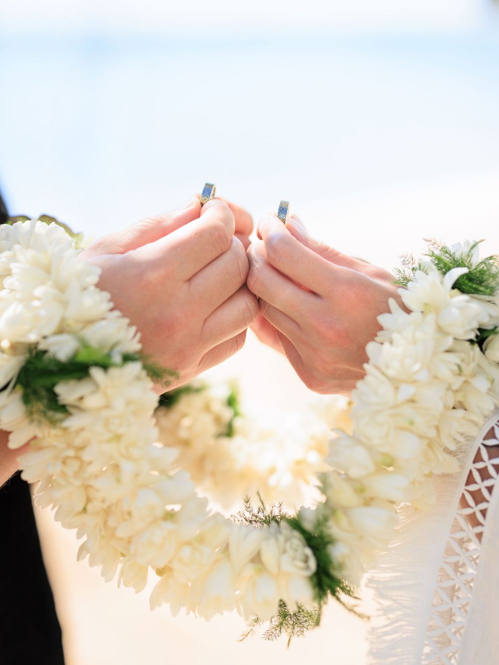 Two hands wearing wedding rings hold white floral leis at a beach wedding ceremony.