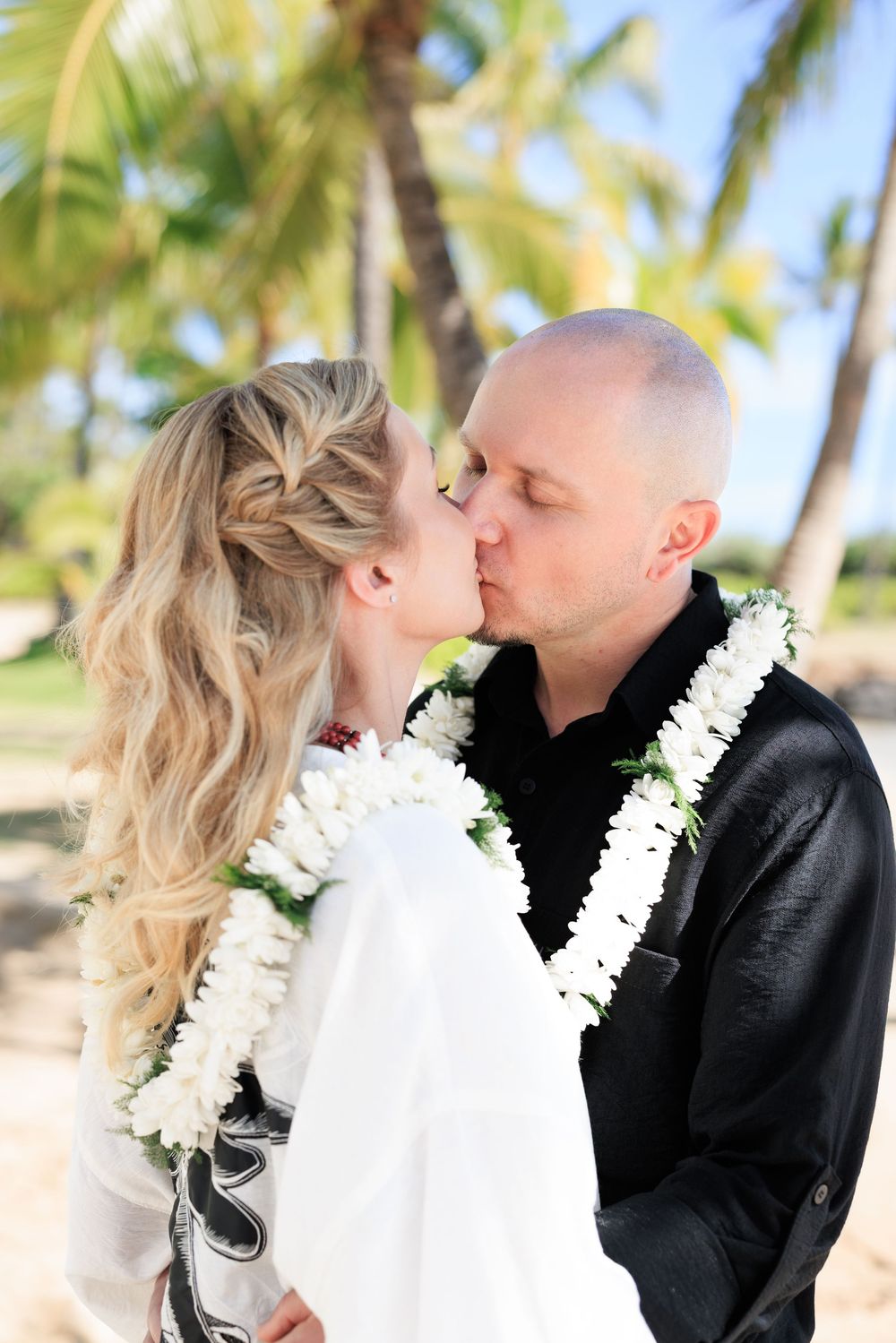 Couple sharing a kiss on a tropical beach, wearing white floral leis, with palm trees in the background.