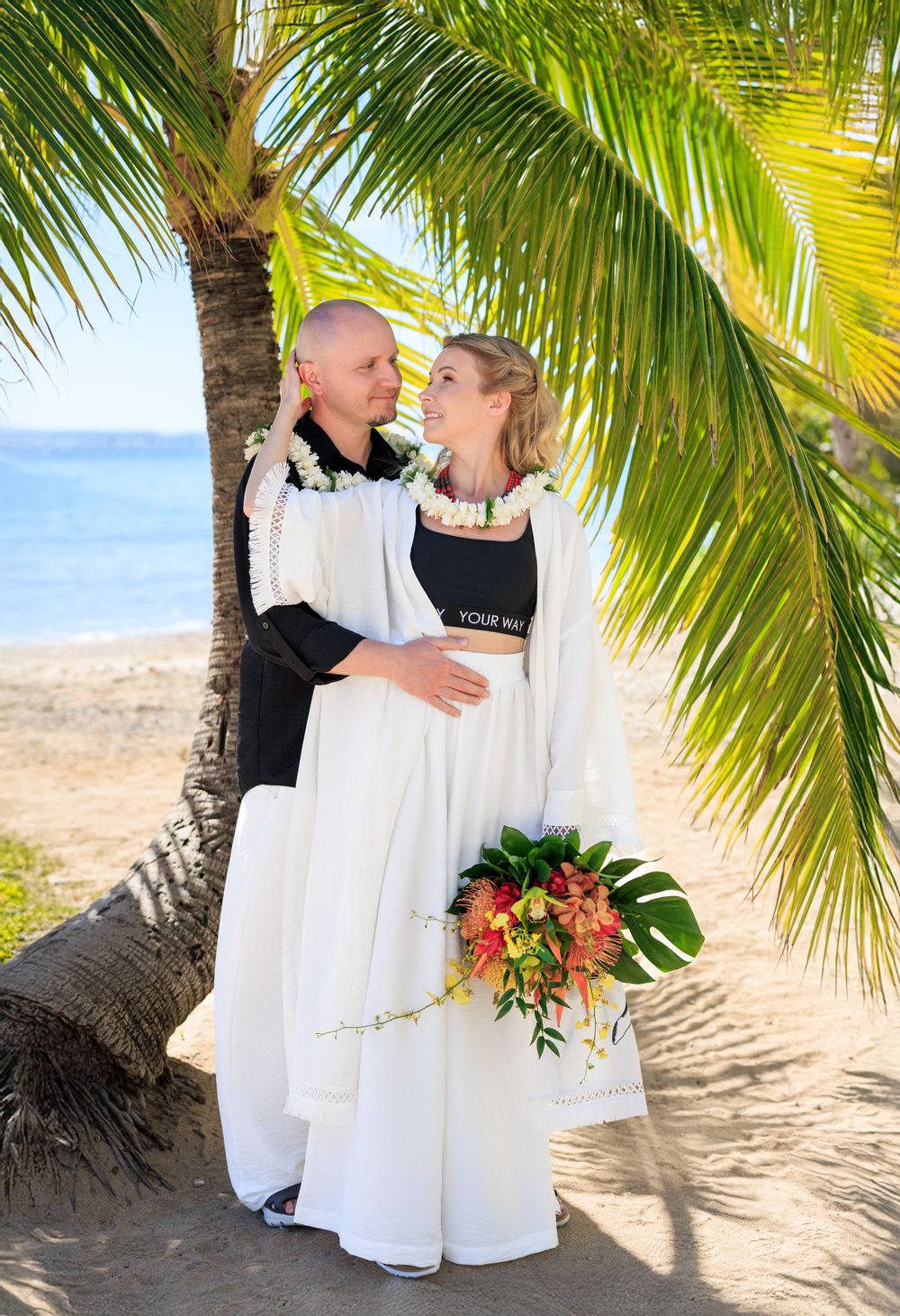 Couple in wedding attire with leis embracing under palm tree on tropical beach, bride holding colorful bouquet.