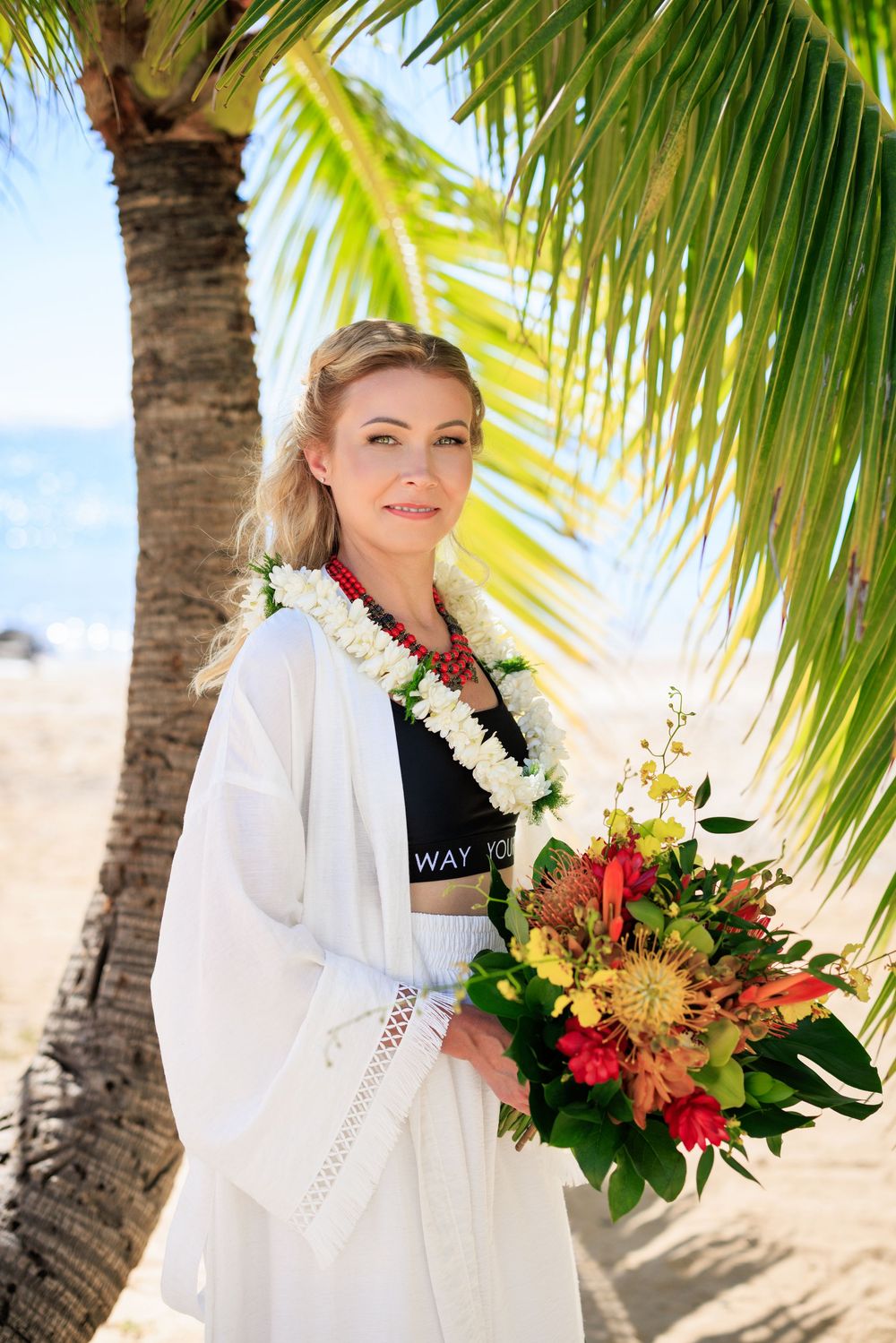 Woman in white wrap holding tropical bouquet with floral lei under palm tree on sunny Hawaiian beach.