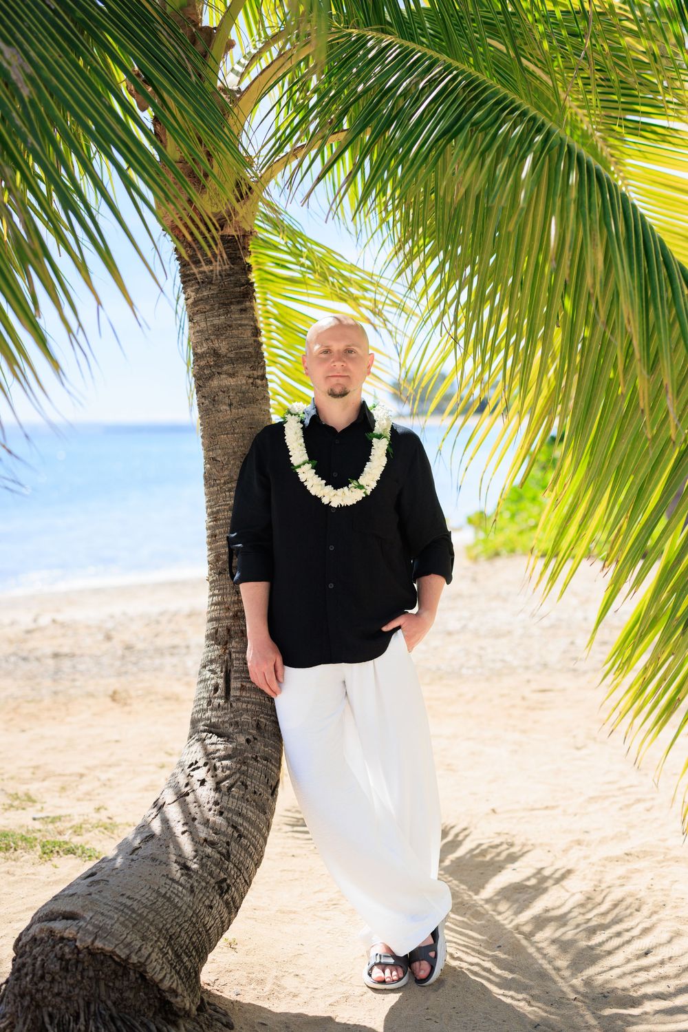 Man in black shirt and white pants wearing a floral lei, leaning against a palm tree on a tropical beach.