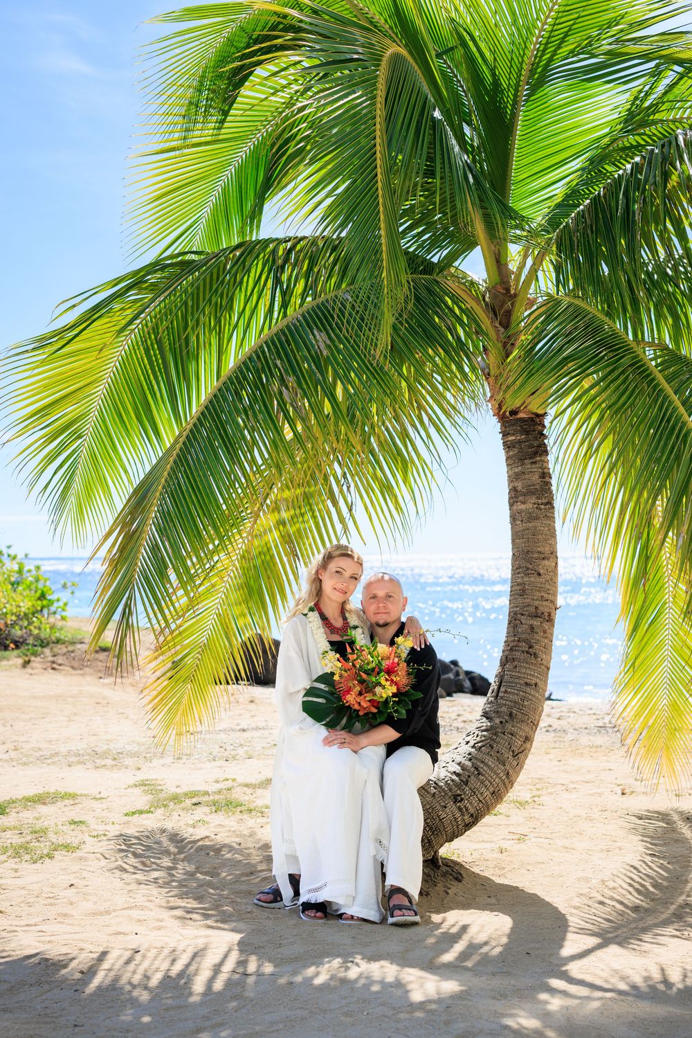 Couple in wedding attire embracing under a tropical palm tree on a sandy beach with ocean views.