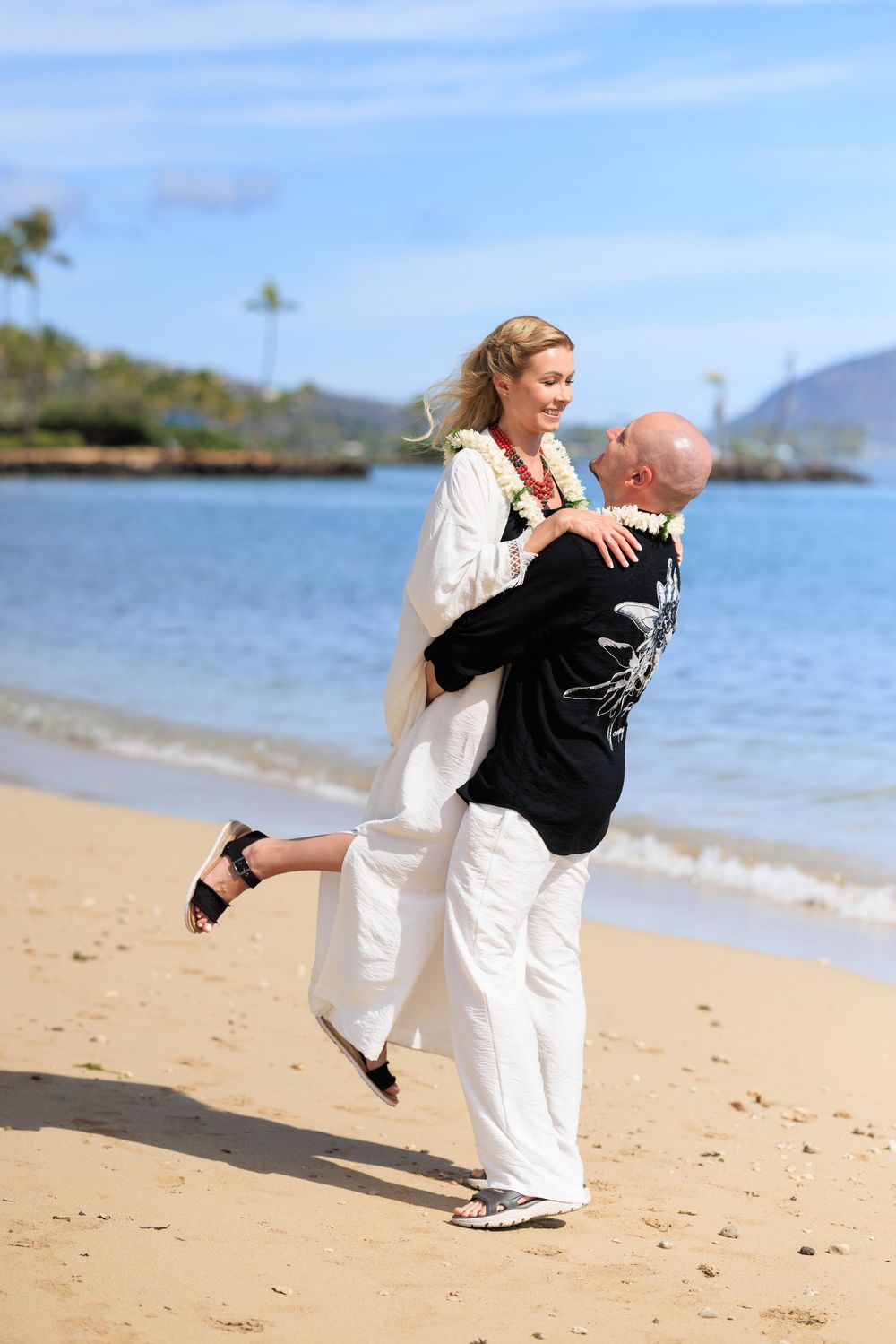 Couple celebrating beach wedding in Hawaii, groom lifting bride in white dress with floral lei near ocean.