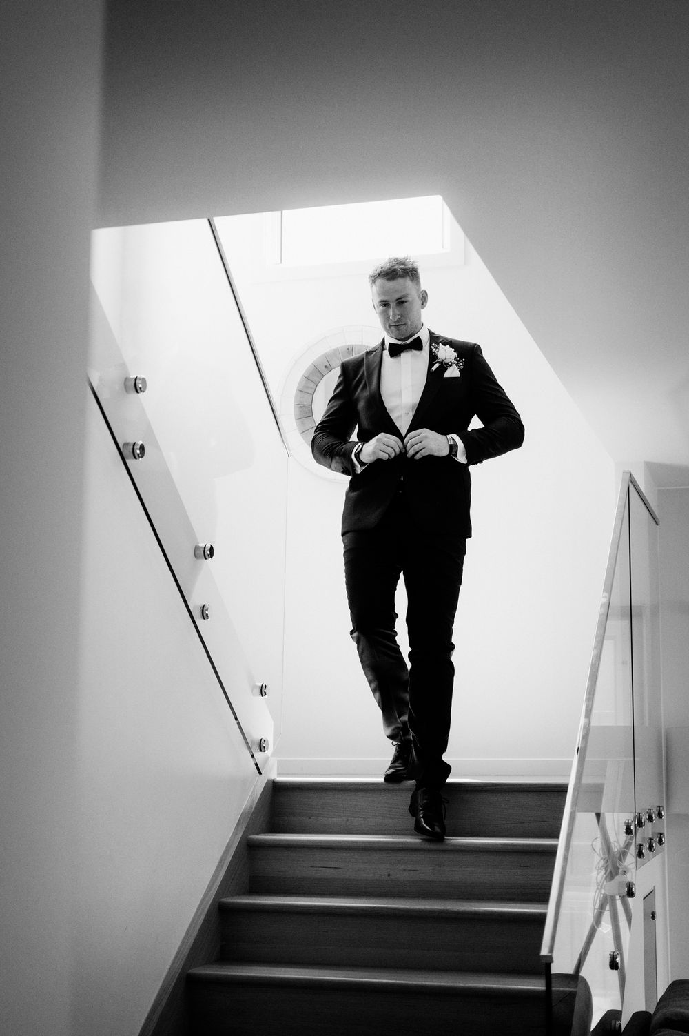 Black and white photo of groom descending modern staircase in classic tuxedo with boutonniere.
