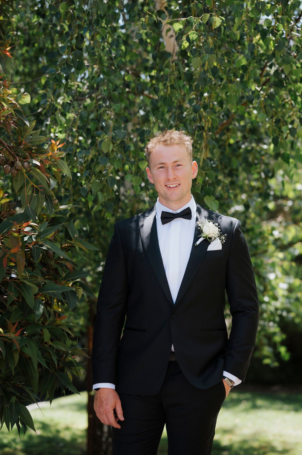 Groom in black tuxedo with bow tie poses in garden setting.