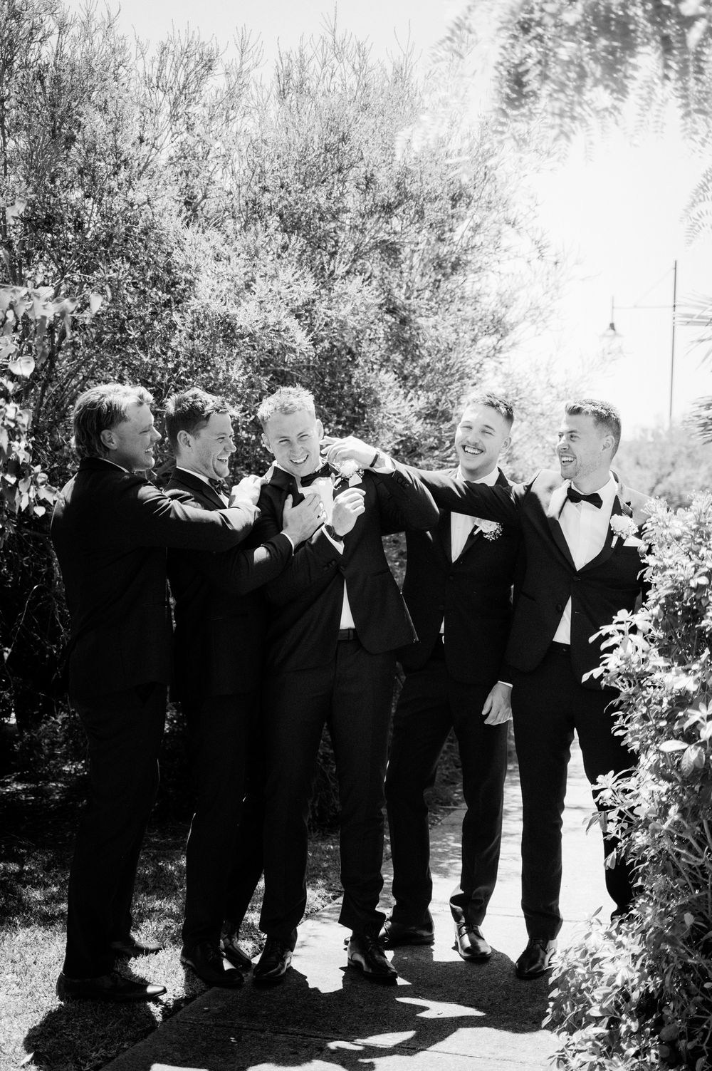 Groomsmen share a playful moment in black and white photo outdoors.