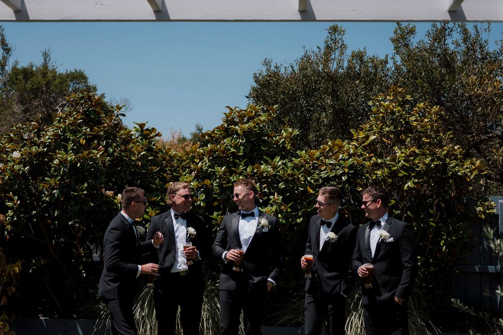Groomsmen in formal black tuxedos gathered outdoors with greenery backdrop.