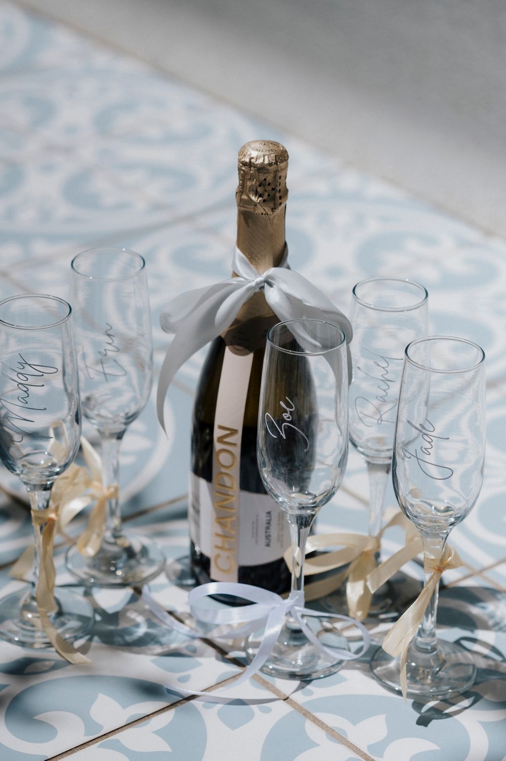Champagne bottle with white bow surrounded by elegant glasses on patterned tablecloth.