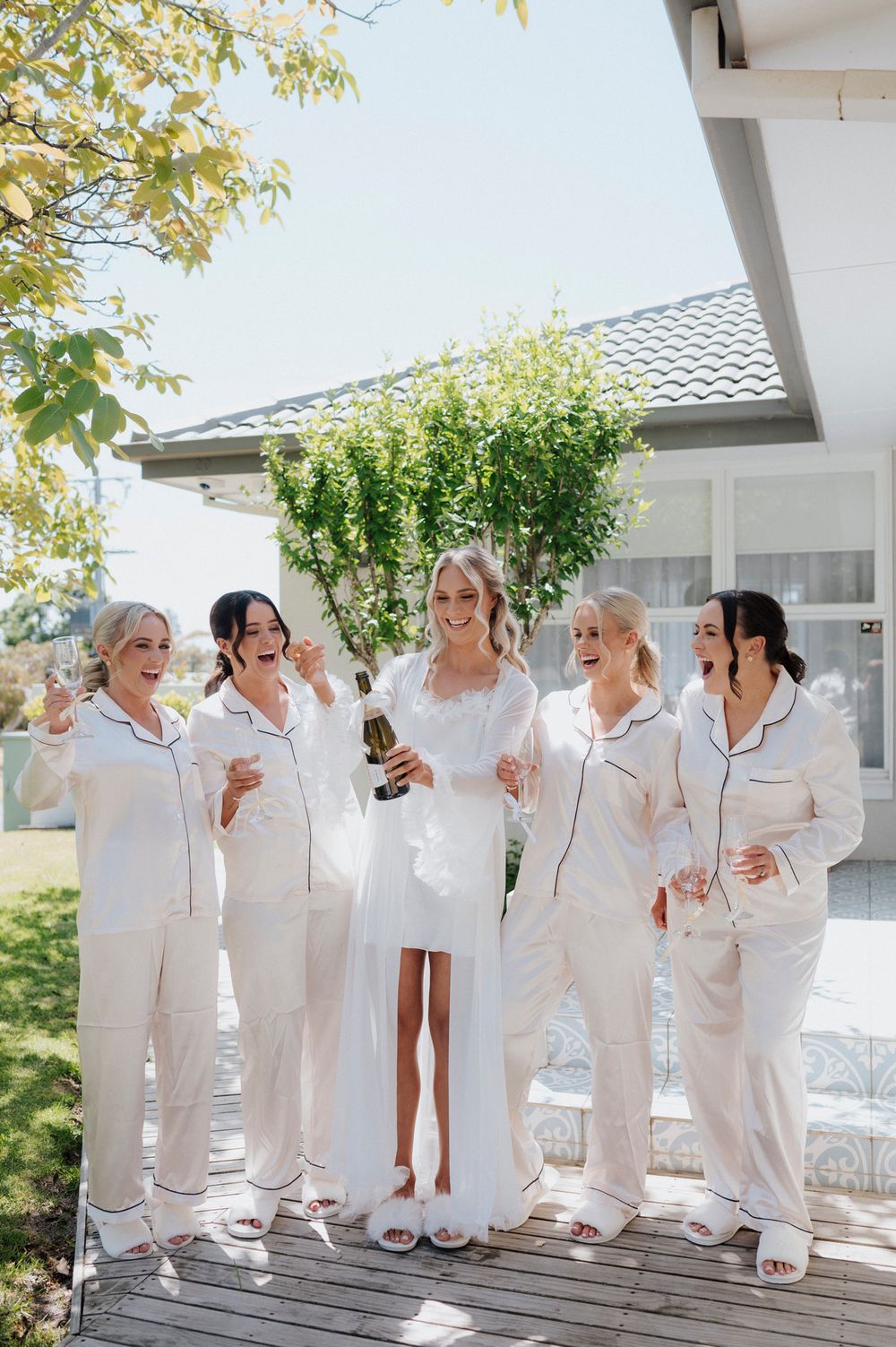 Wedding party in matching white robes celebrating with champagne on outdoor deck.