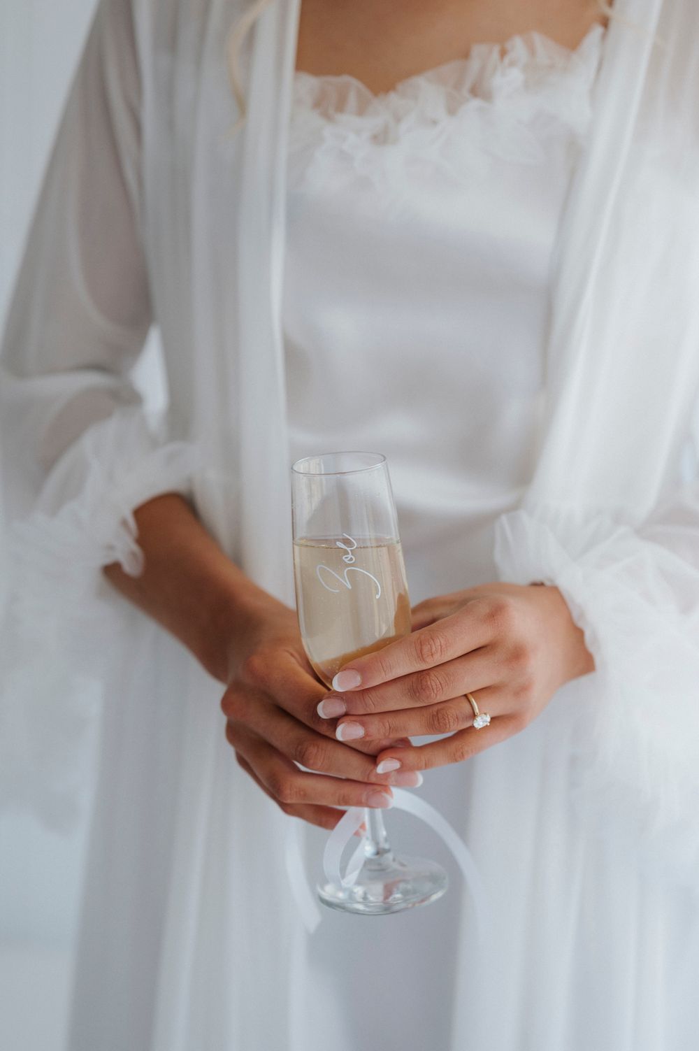 Close-up of hands holding champagne glass in white bridal robe.